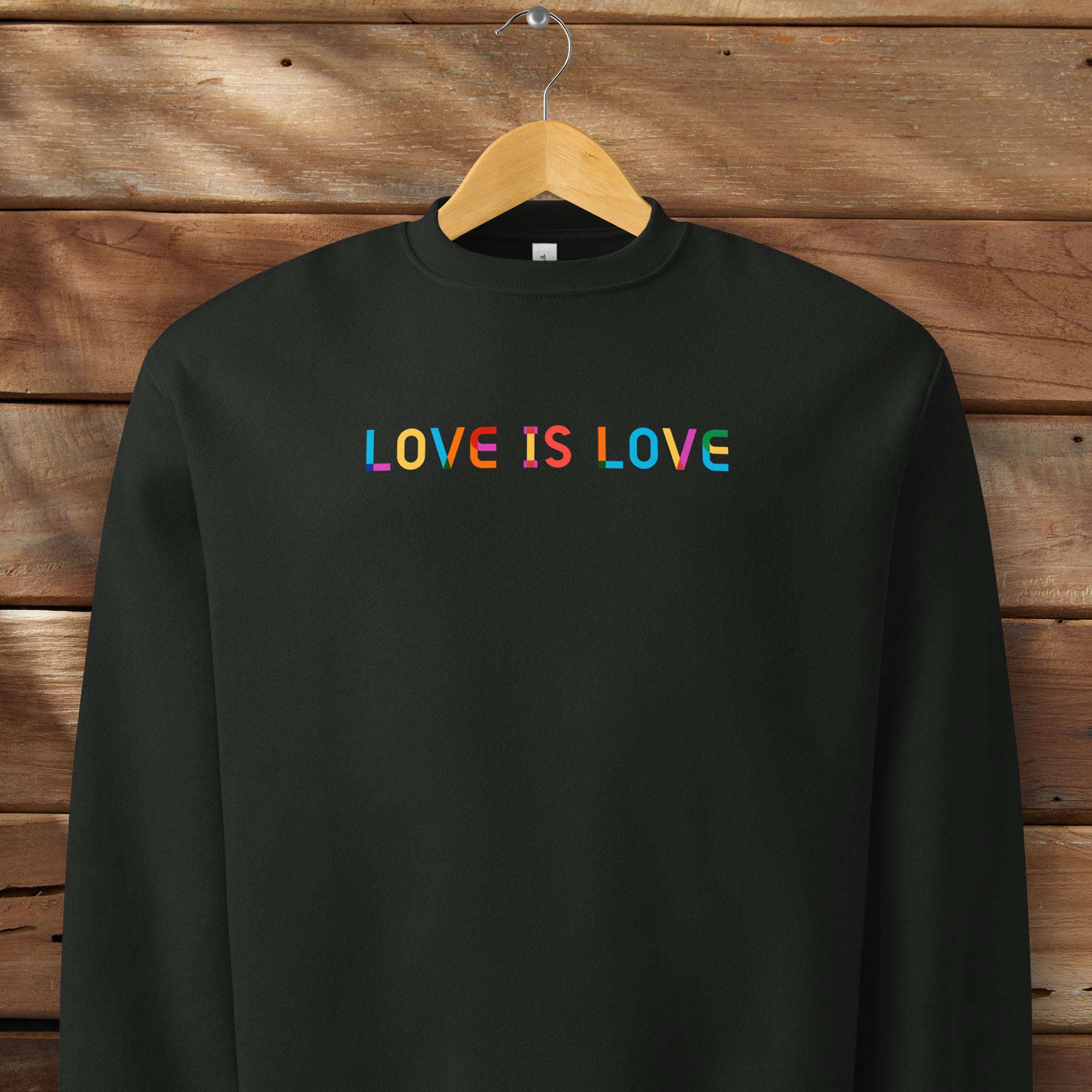 Hanging black Bella+Canvas crewneck sweatshirt with rainbow “love is love” text designed in the Gilbert font.