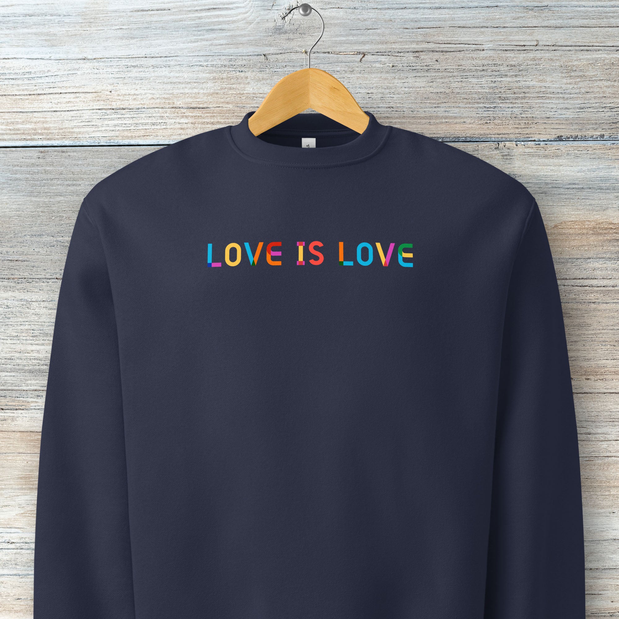 Hanging navy Bella+Canvas crewneck sweatshirt with rainbow “love is love” text designed in the Gilbert font.