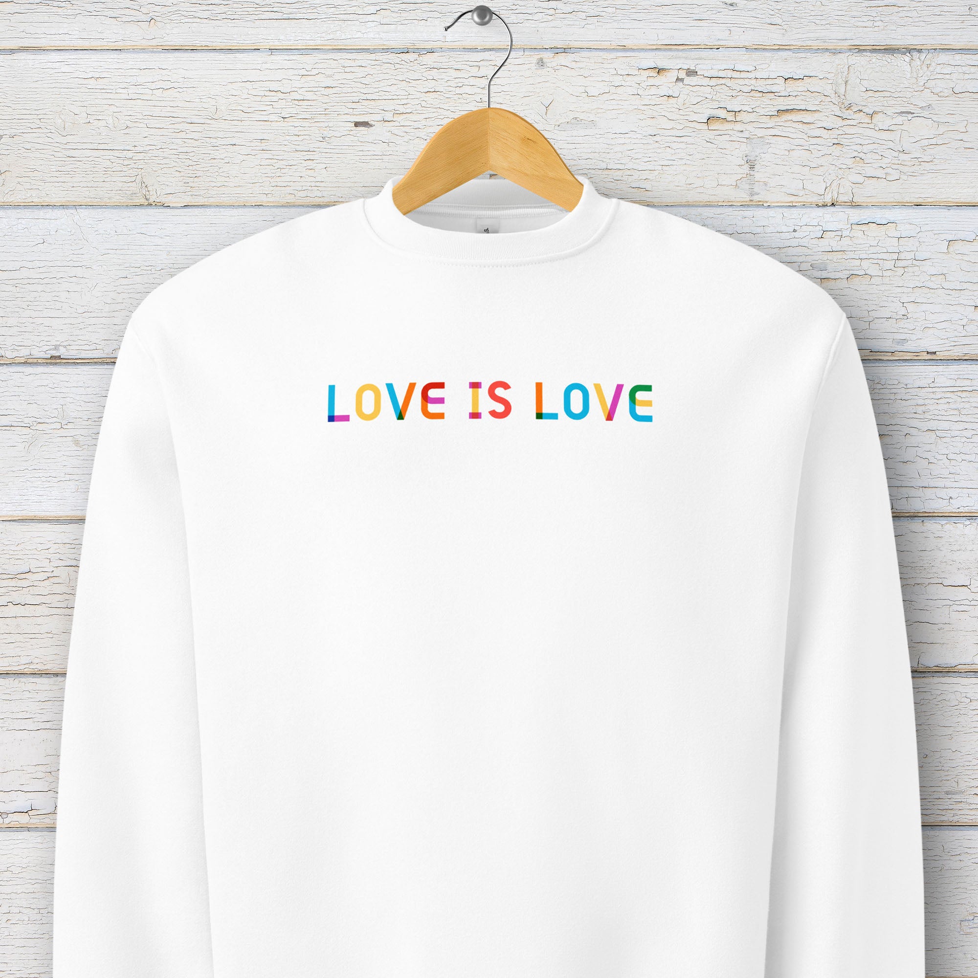 Hanging white Bella+Canvas crewneck sweatshirt with rainbow “love is love” text designed in the Gilbert font.