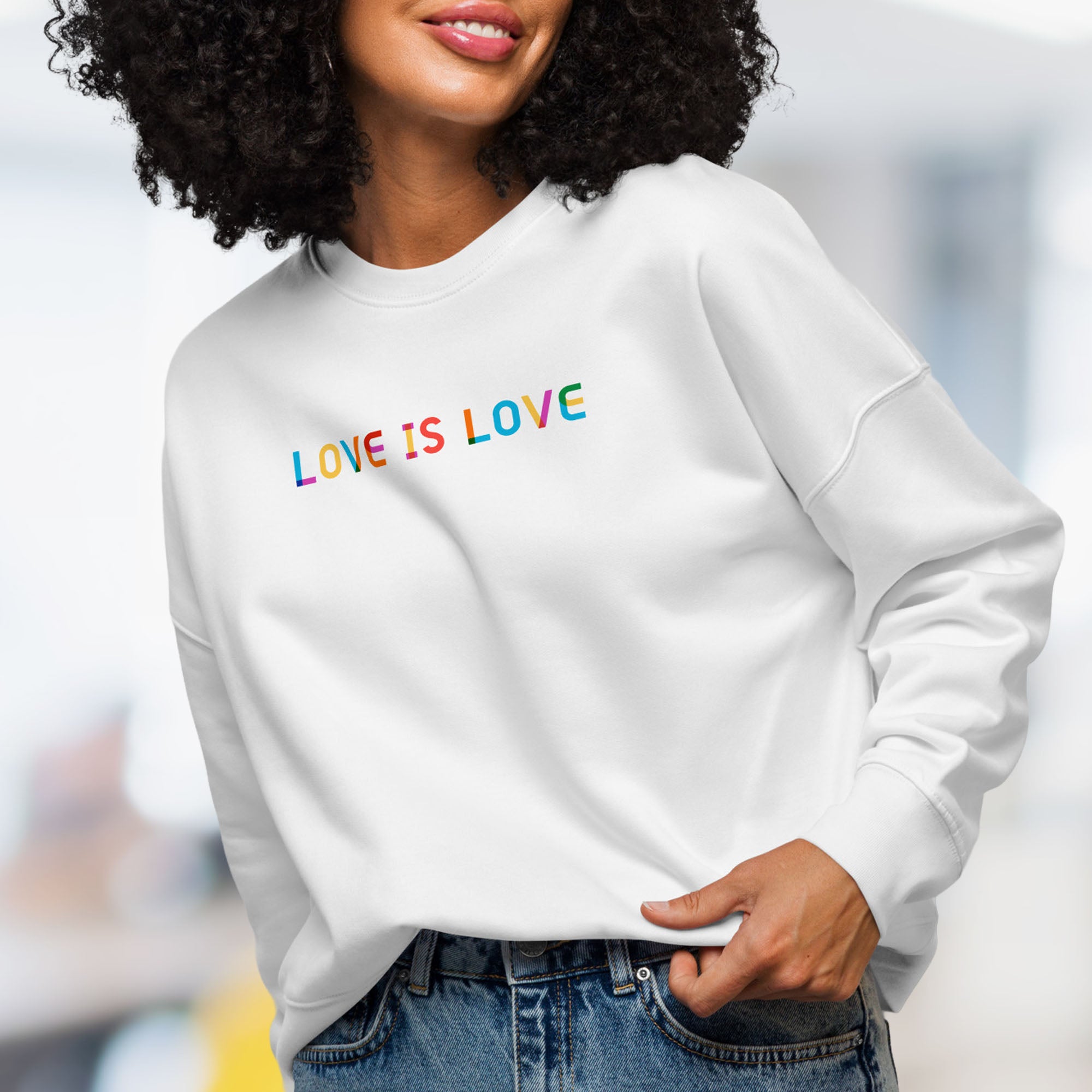 Female model wearing white women's crewneck sweatshirt with rainbow “love is love” text designed in the Gilbert font.