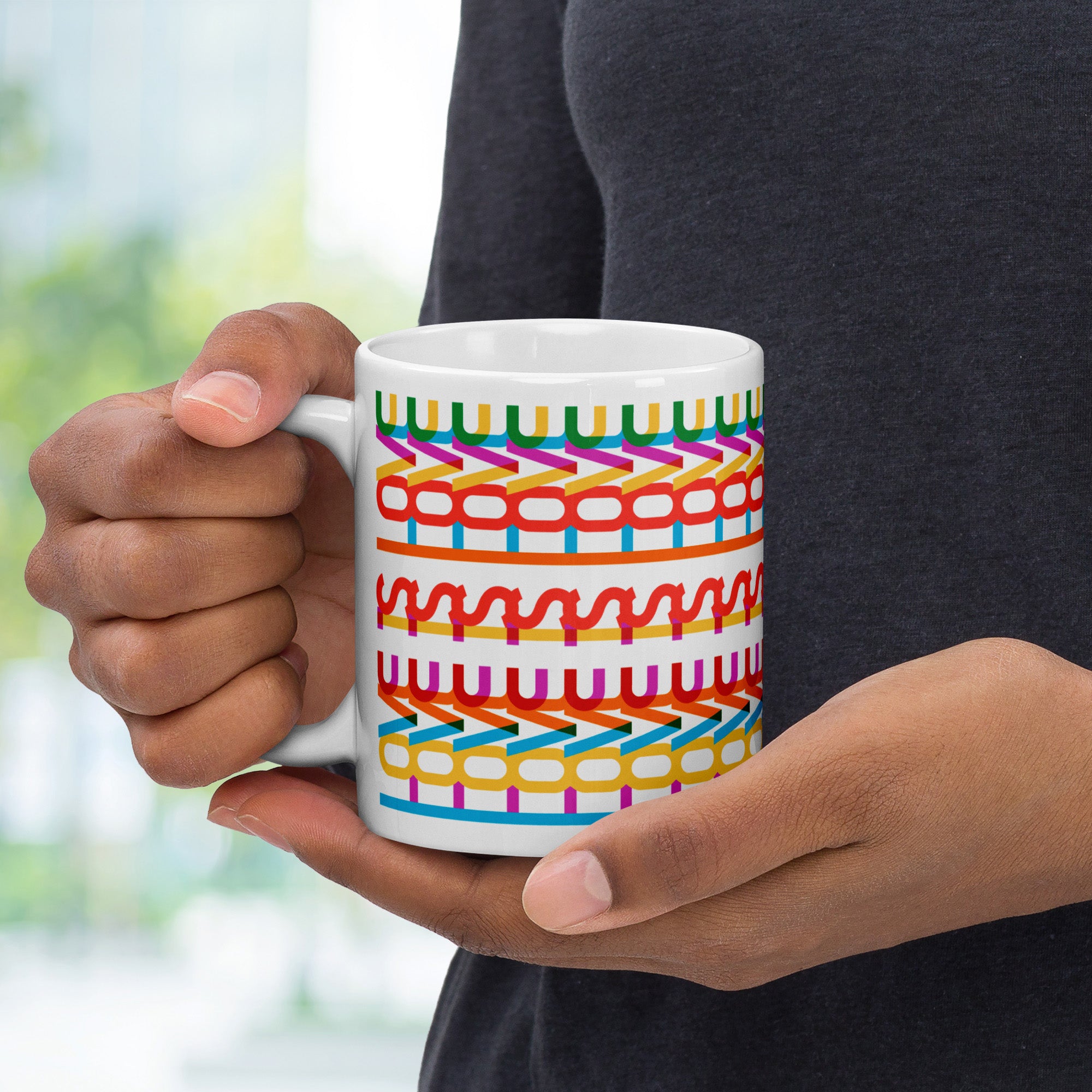 Person holding colorful coffee mug with a repeating hidden “love is love” message in the Gilbert font, featuring bright rainbow-inspired patterns wrapped around the mug.