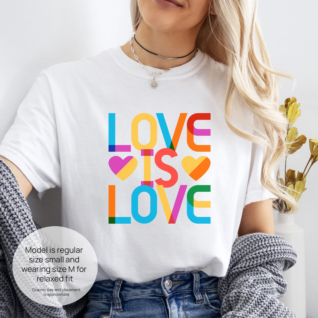 Female model wearing white t-shirt with rainbow “love is love” text block. Includes size and fit information for the model who is regular size small, and wearing a size M shirt for a relaxed fit.
