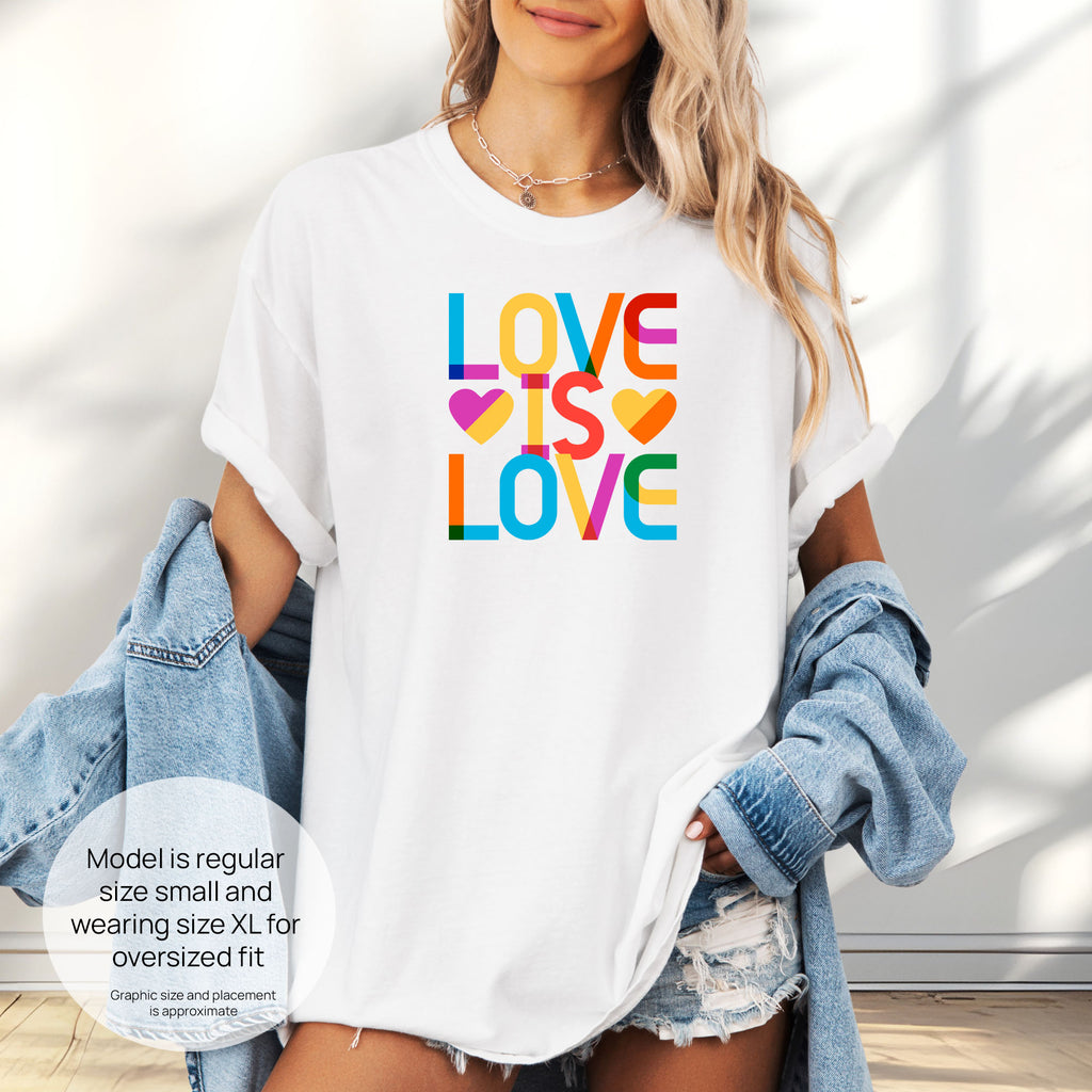 Female model wearing white t-shirt with rainbow “love is love” text block. Includes size and fit information for the model who is regular size small, and wearing a size XL shirt for an oversized fit.
