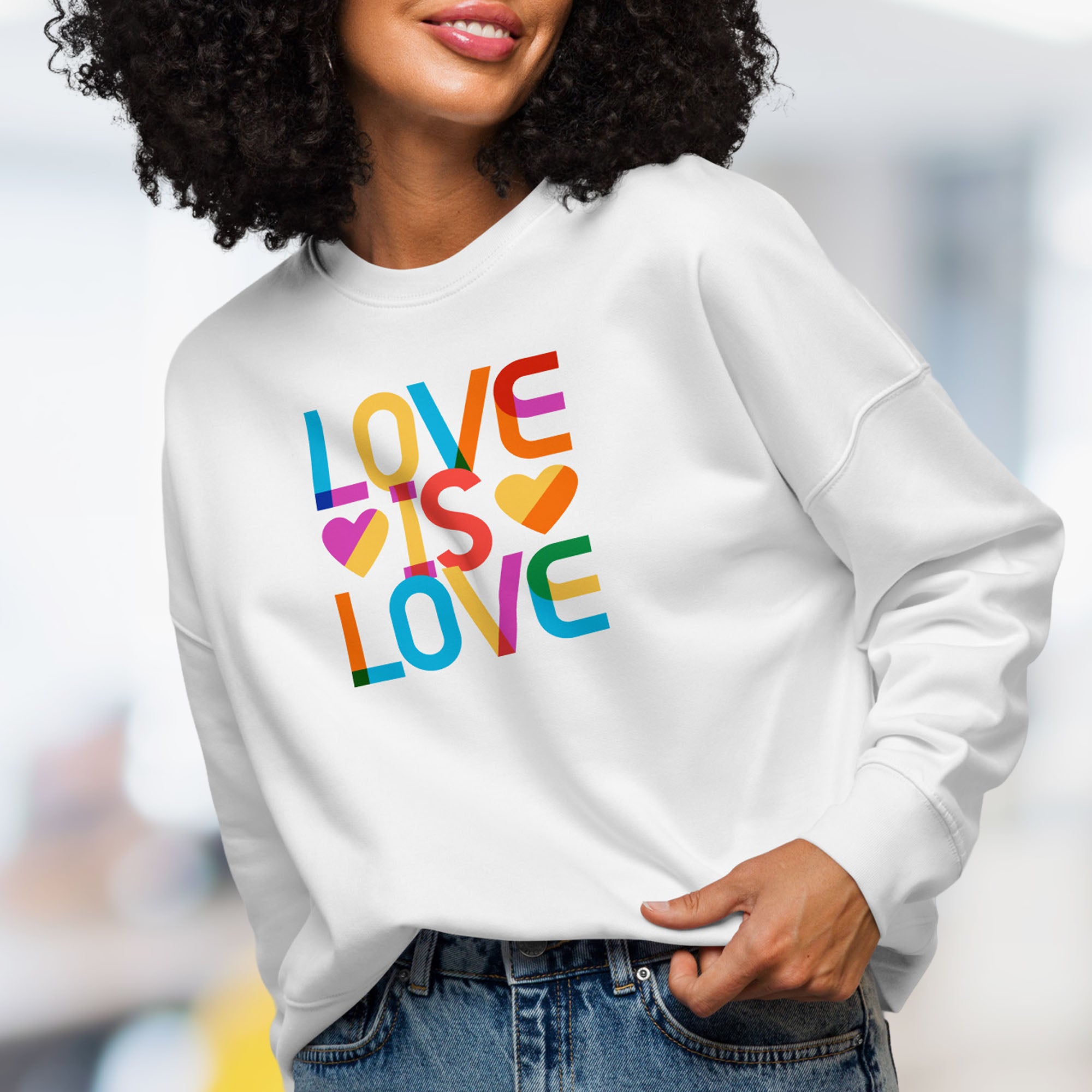Front view of female model wearing white women's sweatshirt with rainbow “love is love” text designed in the Gilbert font.