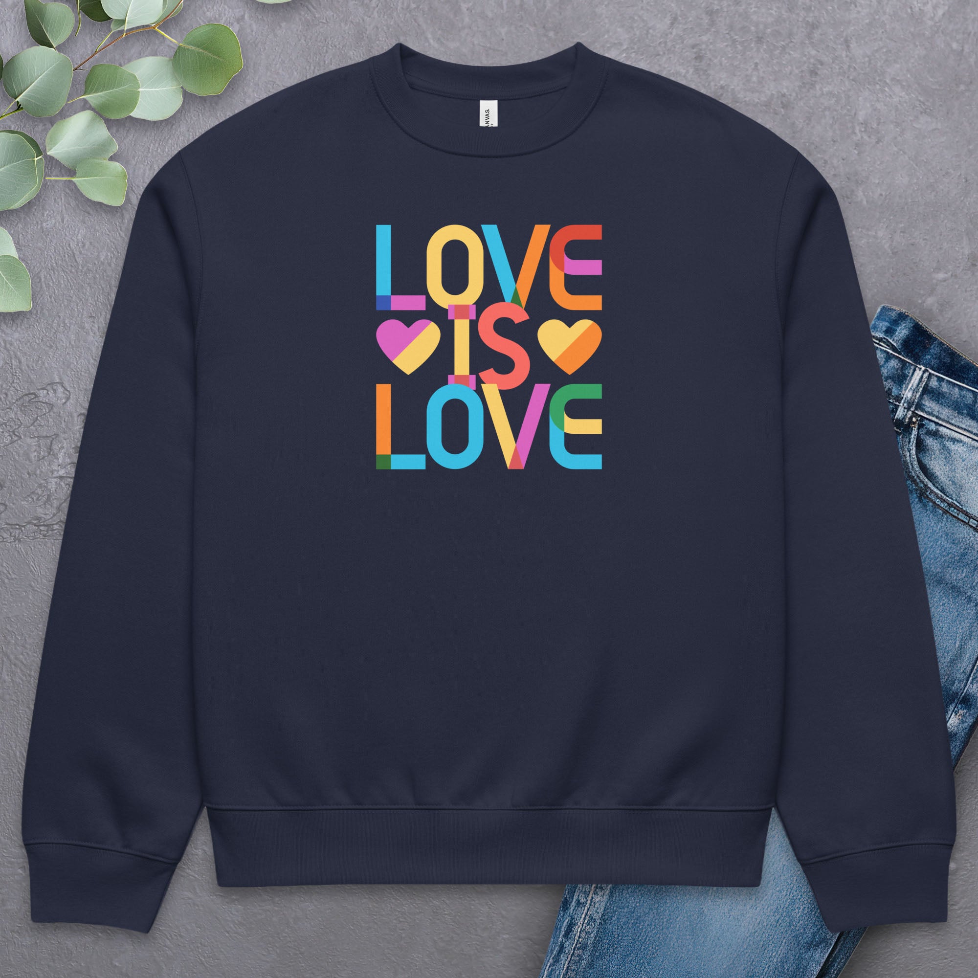 Navy t-shirt with rainbow “love is love” text designed in the Gilbert font, laying flat with a pair of jeans and greenery.