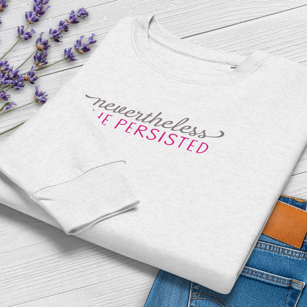 Folded grey crewneck sweatshirt with stylized text graphic that reads "nevertheless she persisted", styled with a pair of jeans and purple flowers.