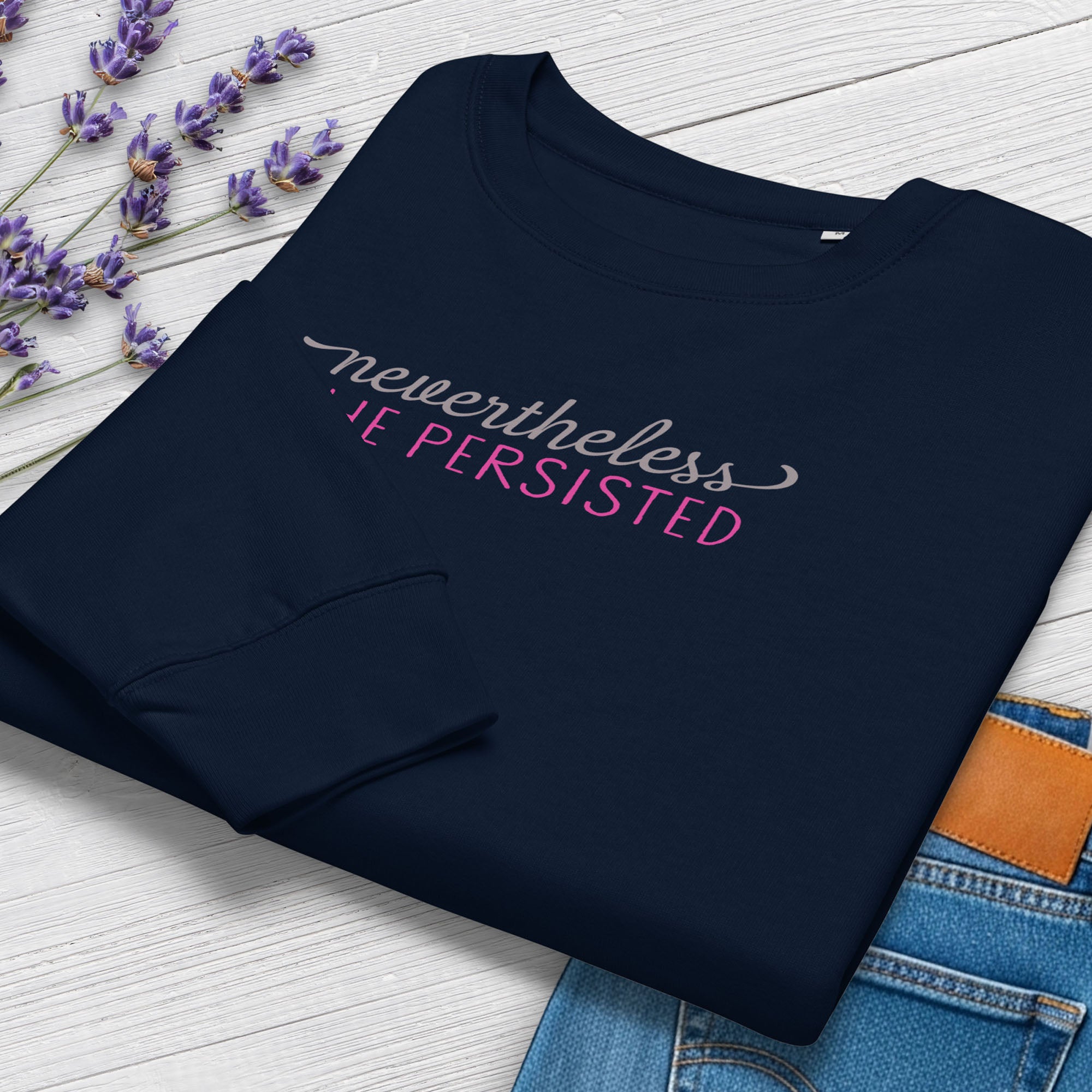 Folded navy crewneck sweatshirt with stylized text graphic that reads "nevertheless she persisted", styled with a pair of jeans and purple flowers.