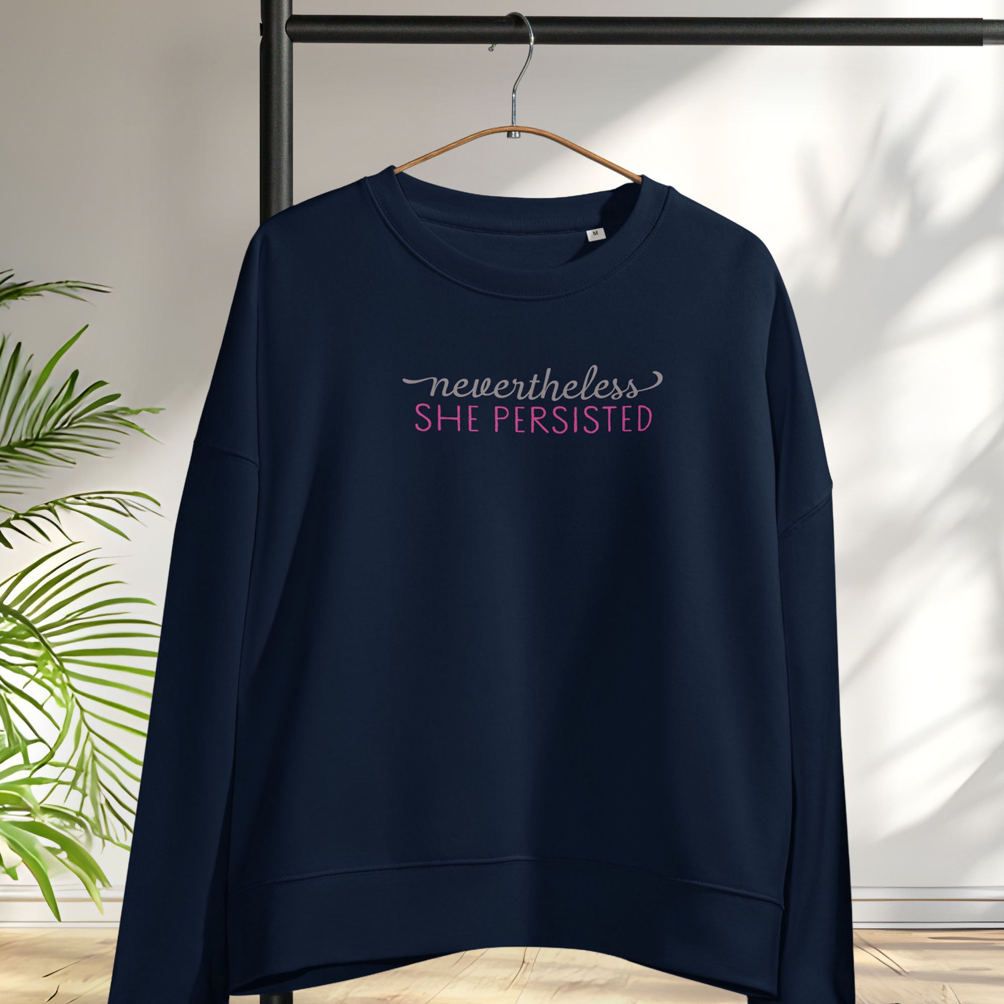 Women's navy sweatshirt with styled text that reads "nevertheless, she persisted", hanging on a hanger.