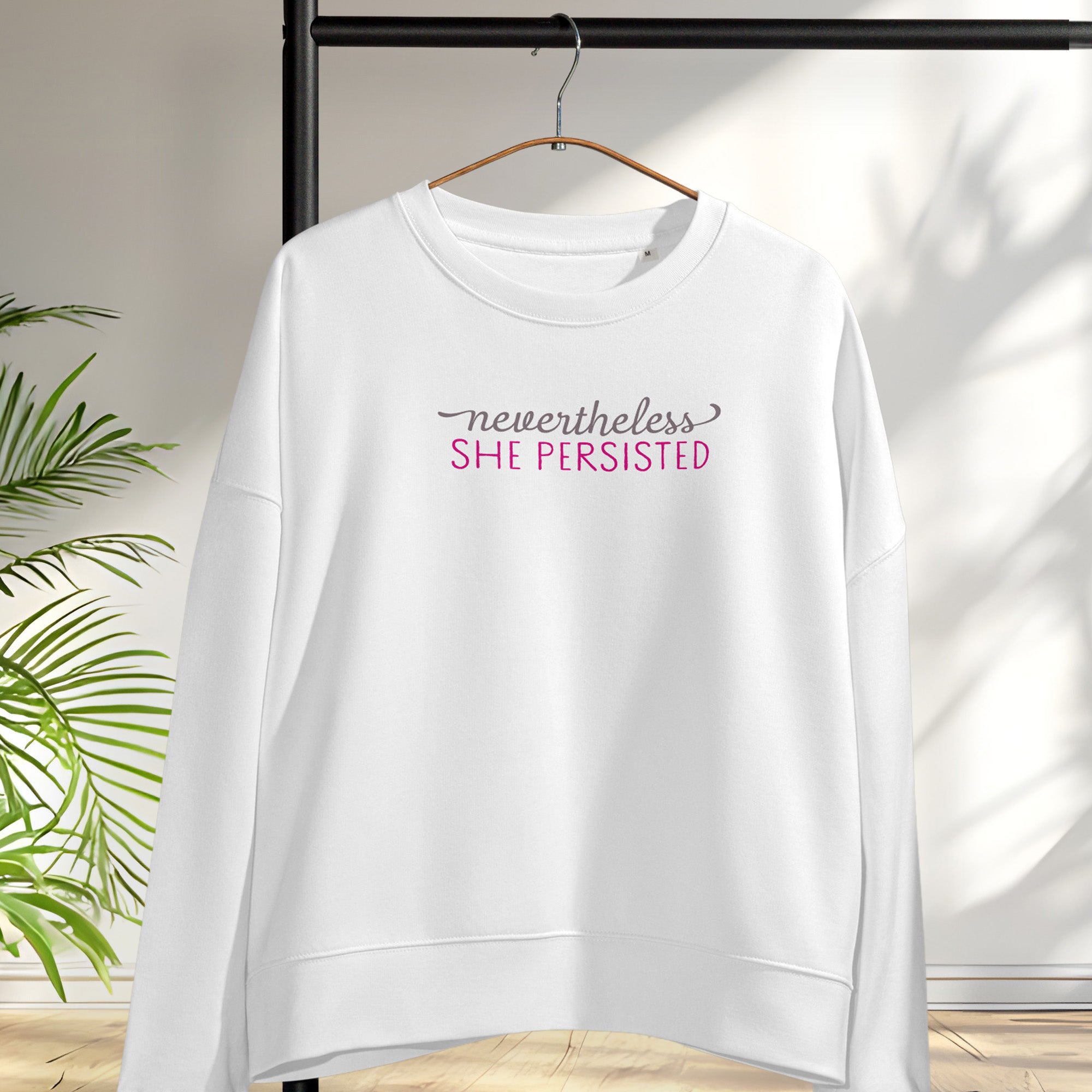 Women's white sweatshirt with styled text that reads "nevertheless, she persisted", hanging on a hanger.
