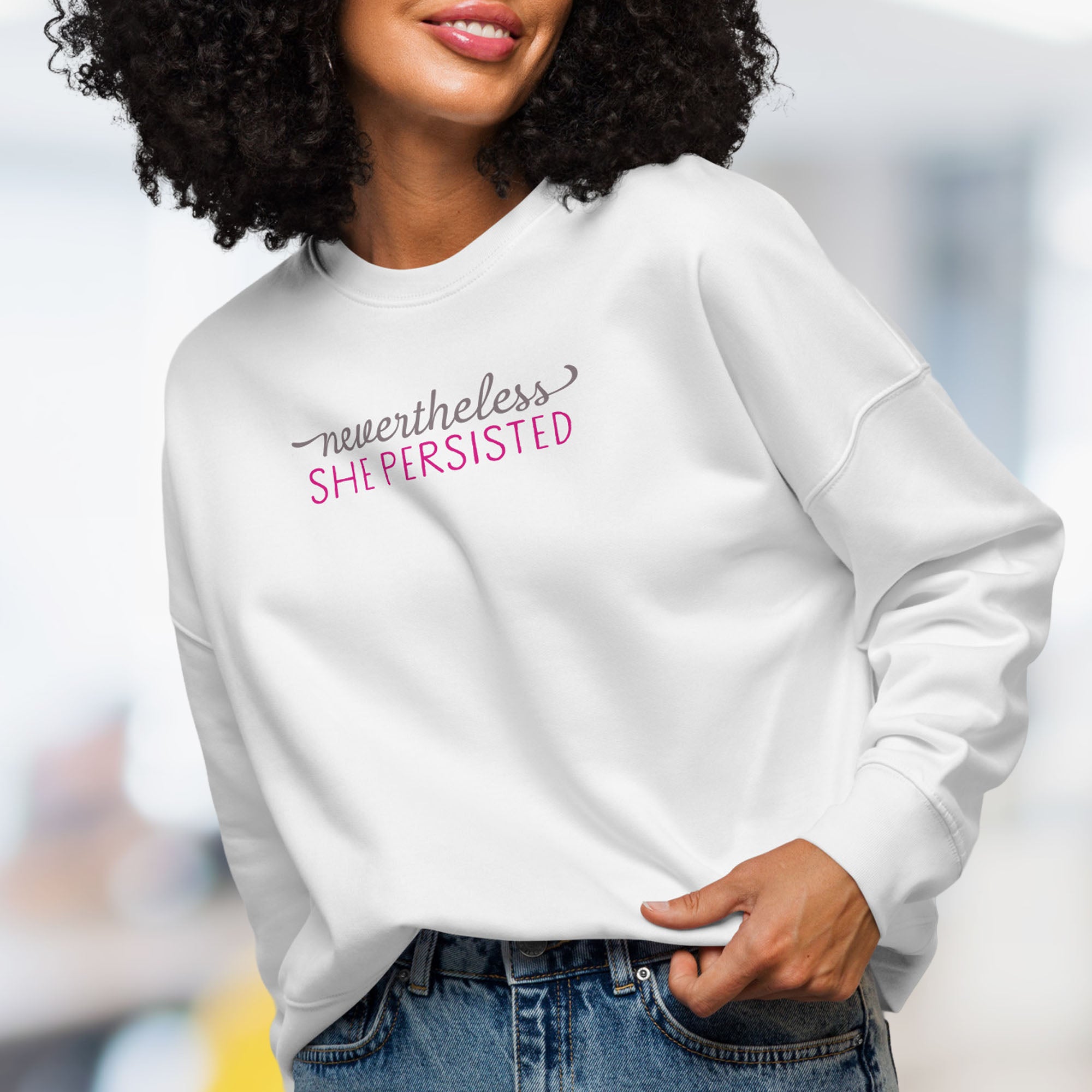 Front view of female model wearing white crewneck sweatshirt with stylized text graphic that reads "nevertheless she persisted".