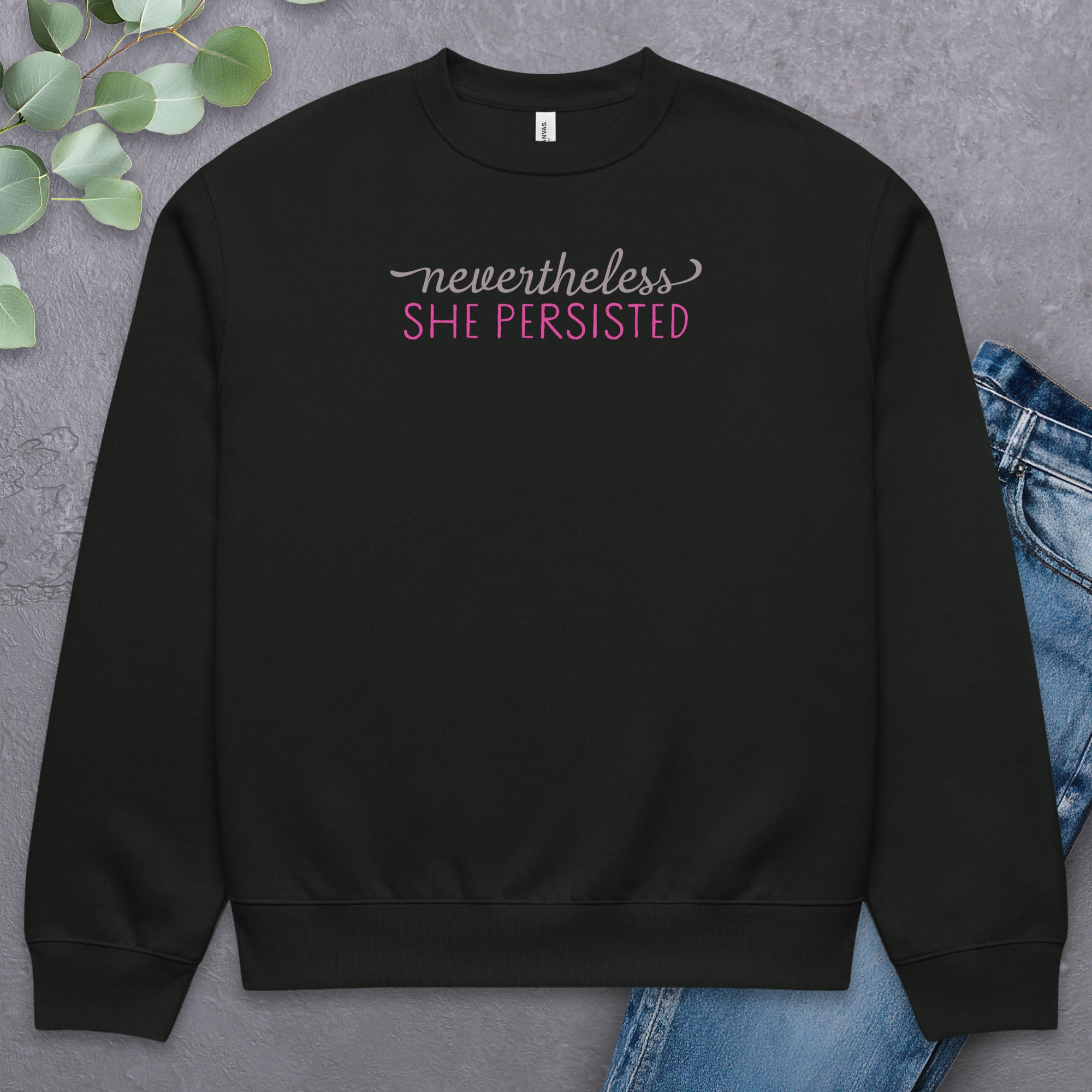 Black sweatshirt with styled text that reads "nevertheless, she persisted", laying flat with a pair of jeans.