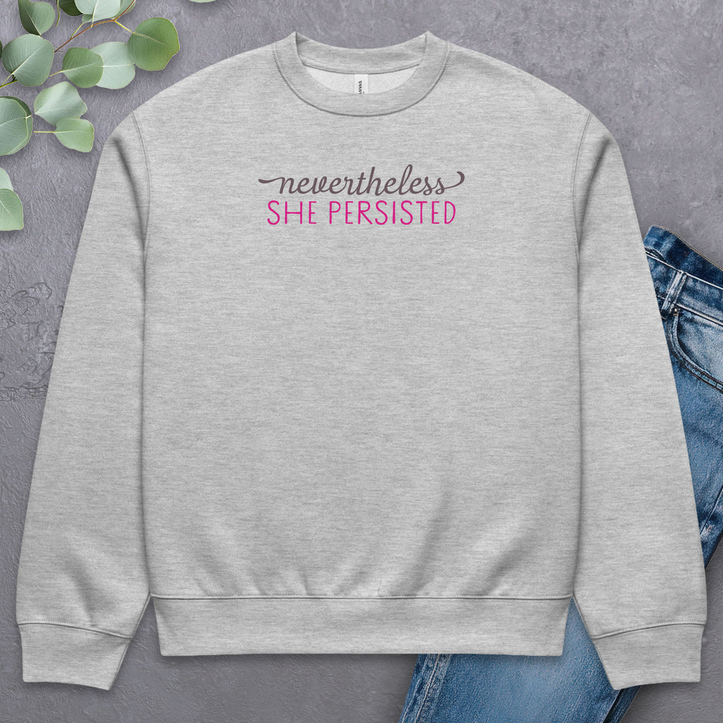 Athletic heather sweatshirt with styled text that reads "nevertheless, she persisted", laying flat with a pair of jeans.
