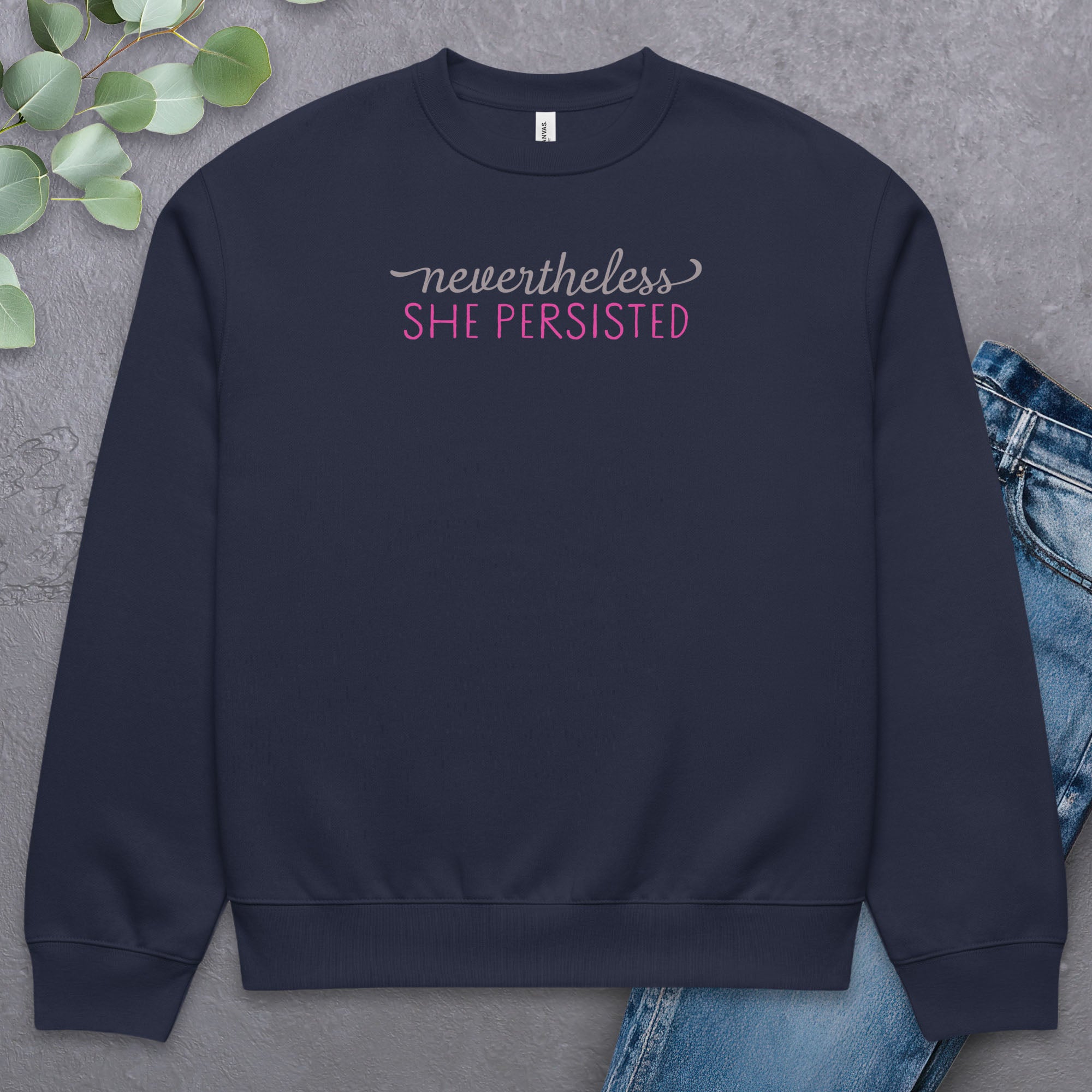 Navy sweatshirt with styled text that reads "nevertheless, she persisted", laying flat with a pair of jeans.