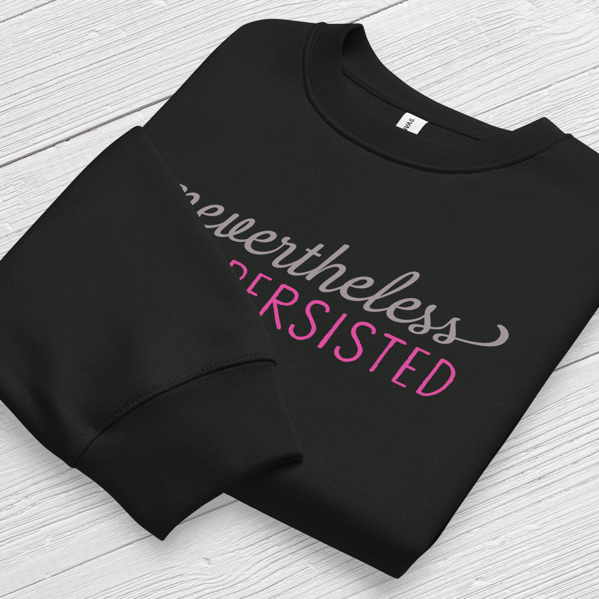 Black sweatshirt with styled text that reads "nevertheless, she persisted", folded flat showing graphic and cuff detail.