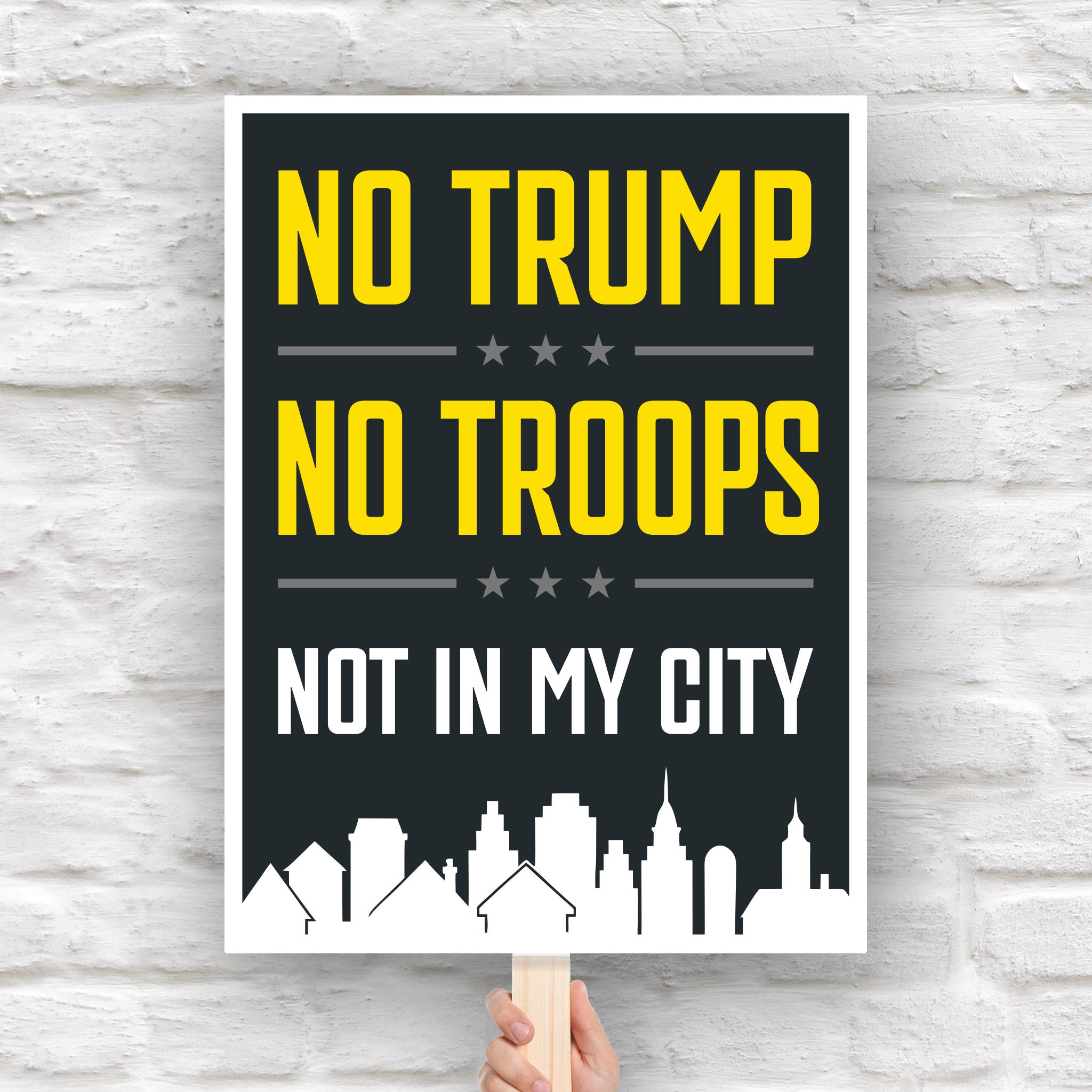 Anti-Trump protest sign with bold yellow and white text reading “No Trump No Troops Not in My City” over a dark background with a city skyline silhouette.