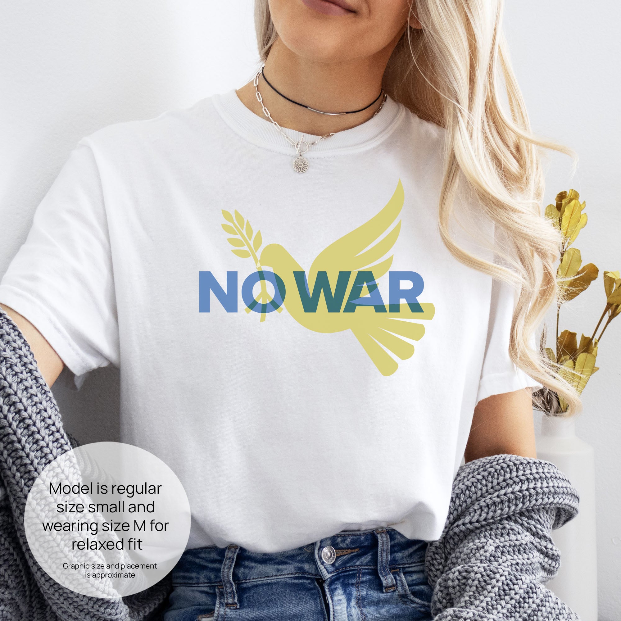 Female model wearing white t-shirt featuring blue “No War” text over a yellow peace dove carrying an olive branch that forks into a peace sign over the text. Includes size and fit information for the model who is regular size small, and wearing a size M shirt for a relaxed fit.
