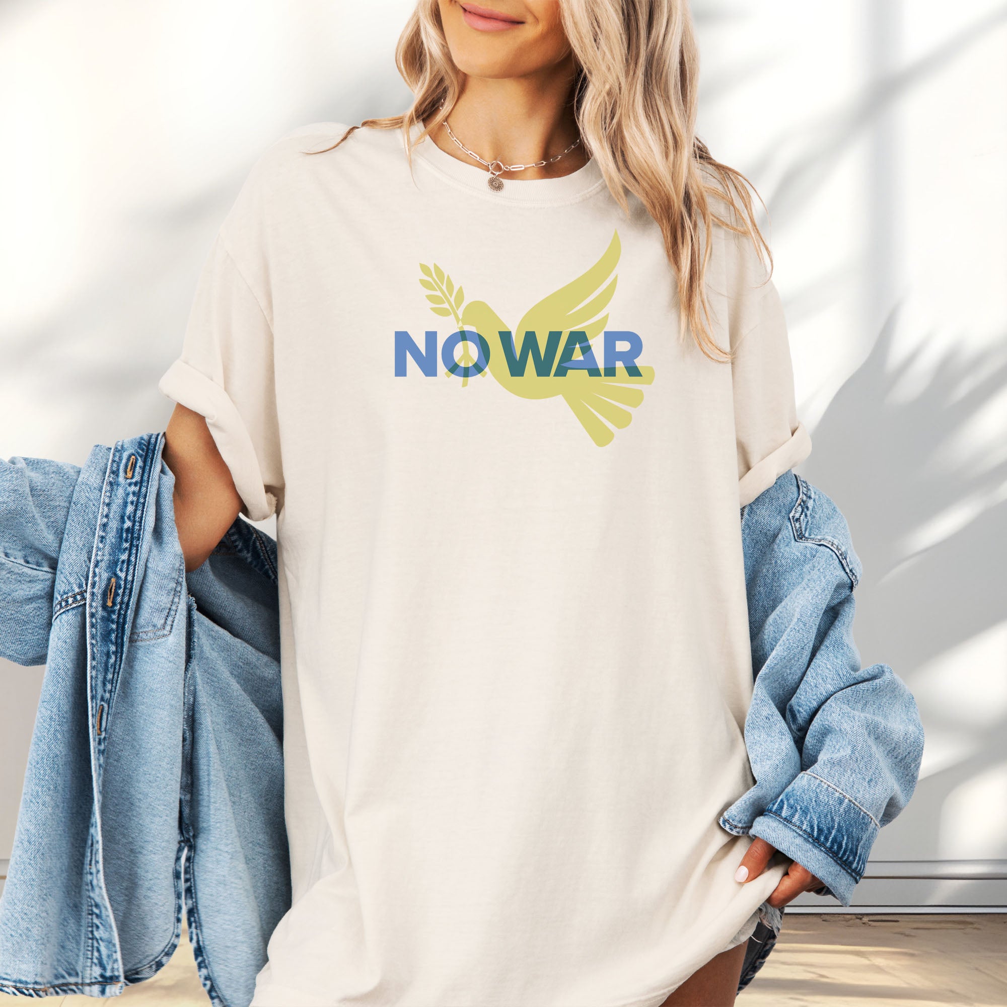 Female model wearing oversized ivory t-shirt featuring blue “No War” text over a yellow peace dove carrying an olive branch that forks into a peace sign over the text..