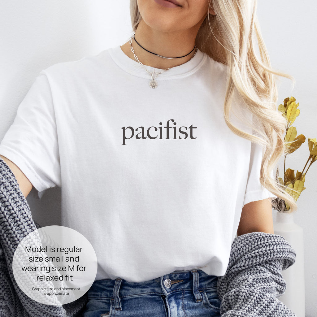 Female model wearing white t-shirt with minimalist serif text reading "pacifist." Includes size and fit information for the model who is regular size small, and wearing a size M shirt for a relaxed fit.
