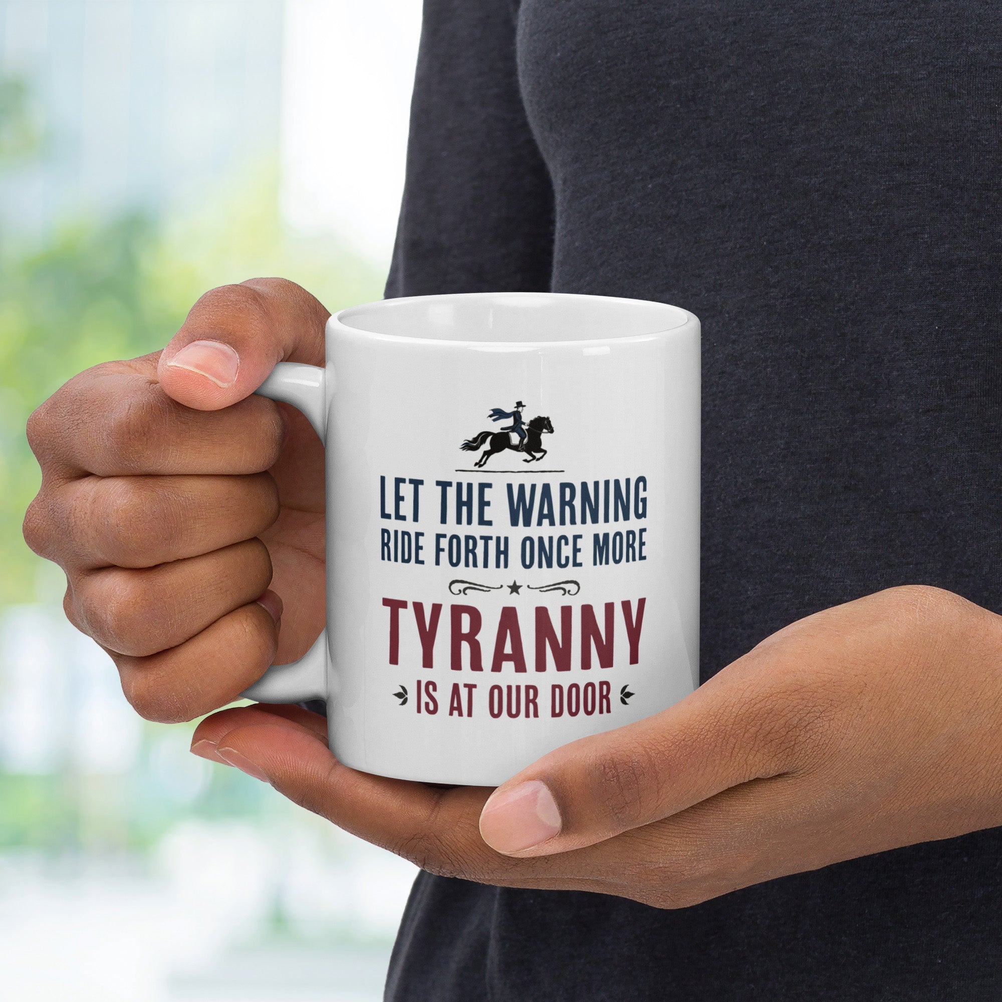 White ceramic coffee mug with a historical-style design reading “Let the warning ride forth once more — Tyranny is at our door,” held by a person with handle on left.