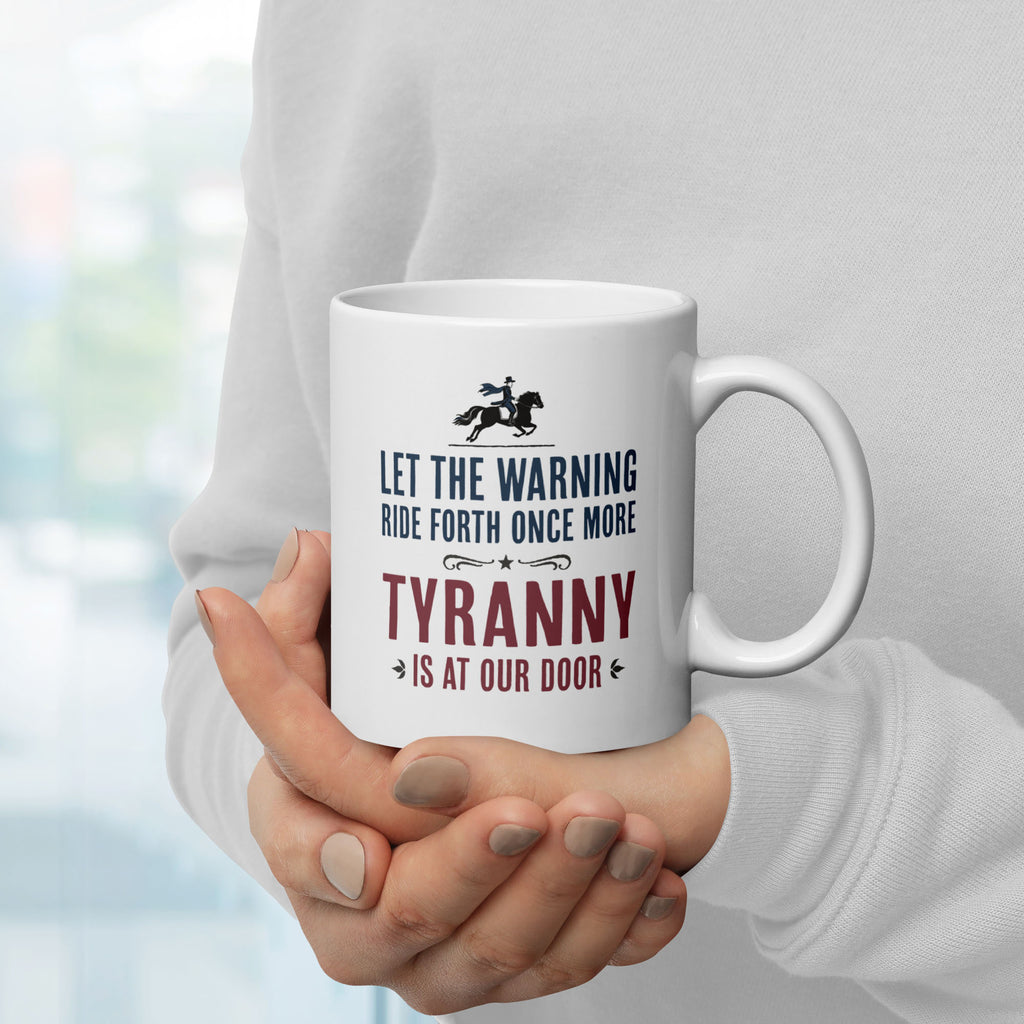 White ceramic coffee mug with a historical-style design reading “Let the warning ride forth once more — Tyranny is at our door,” held by a person with handle on right.
