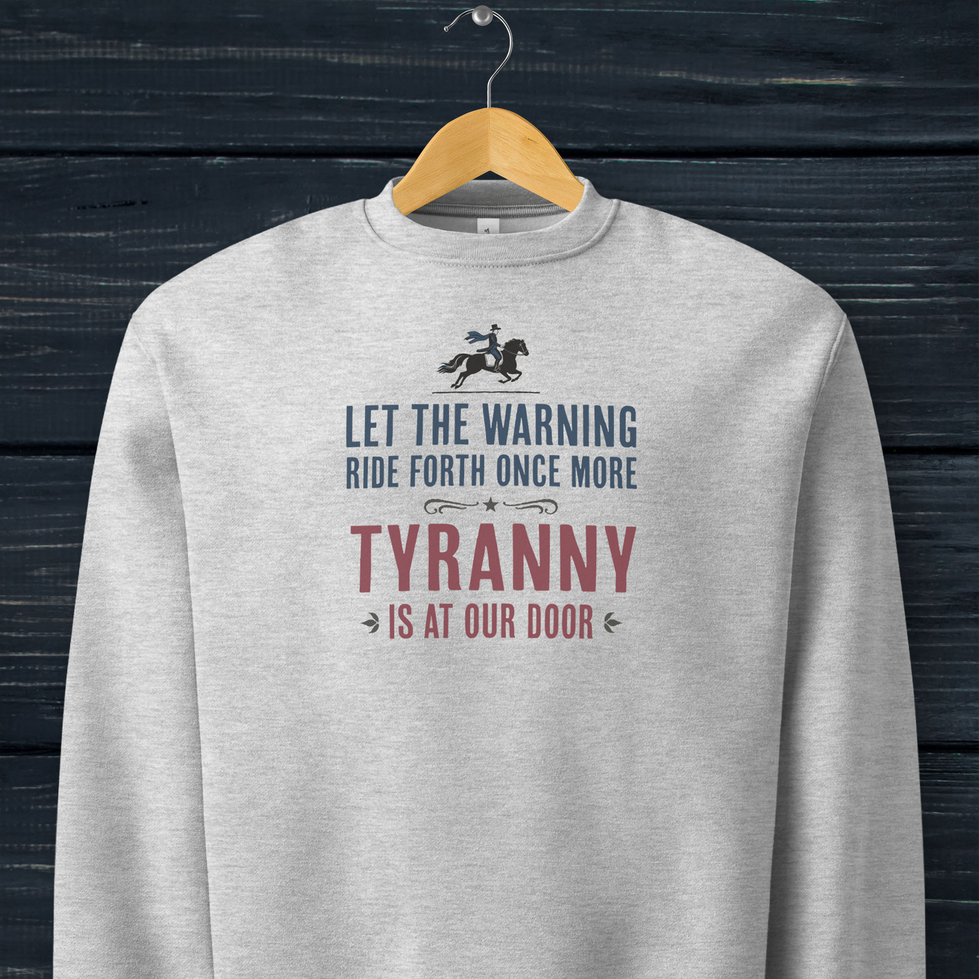 Athletic heather grey sweatshirt with a silhouette of Paul Revere on horseback and the text “Let the warning ride forth once more – Tyranny is at our door” in blue and red, hanging on a hanger.