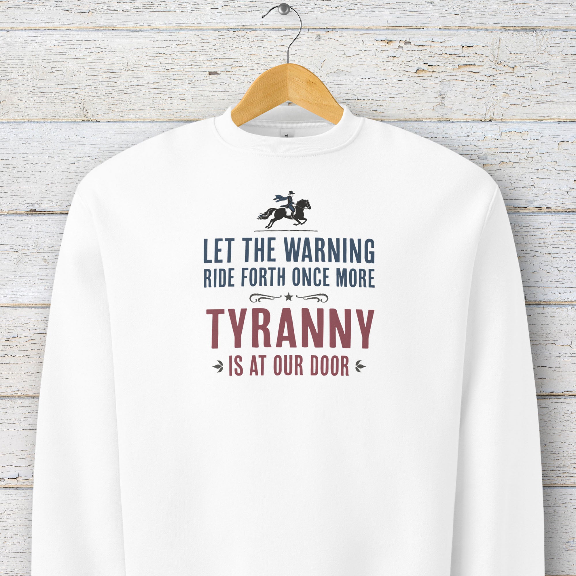 White sweatshirt with a silhouette of Paul Revere on horseback and the text “Let the warning ride forth once more – Tyranny is at our door” in blue and red, hanging on a hanger.
