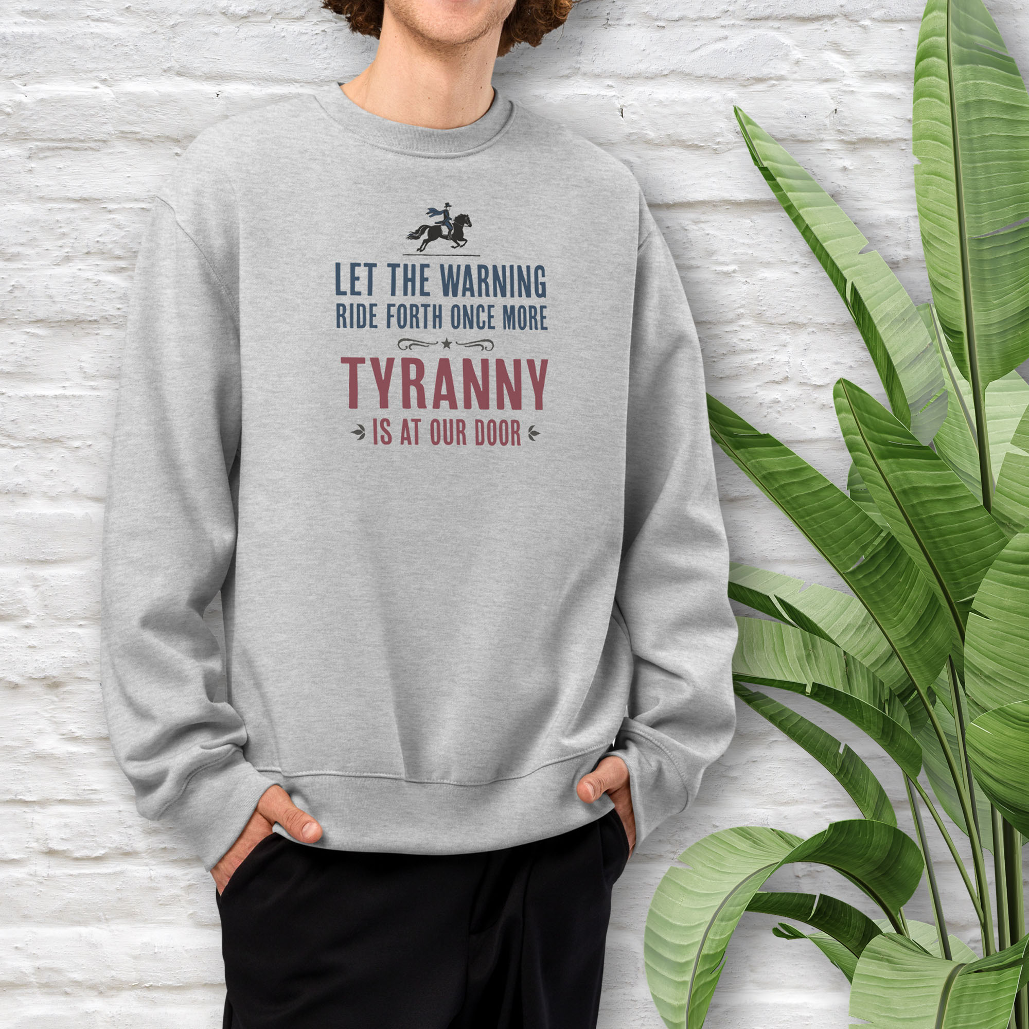 Male model wearing athletic heather grey sweatshirt with a silhouette of Paul Revere on horseback and the text “Let the warning ride forth once more – Tyranny is at our door” in blue and red.