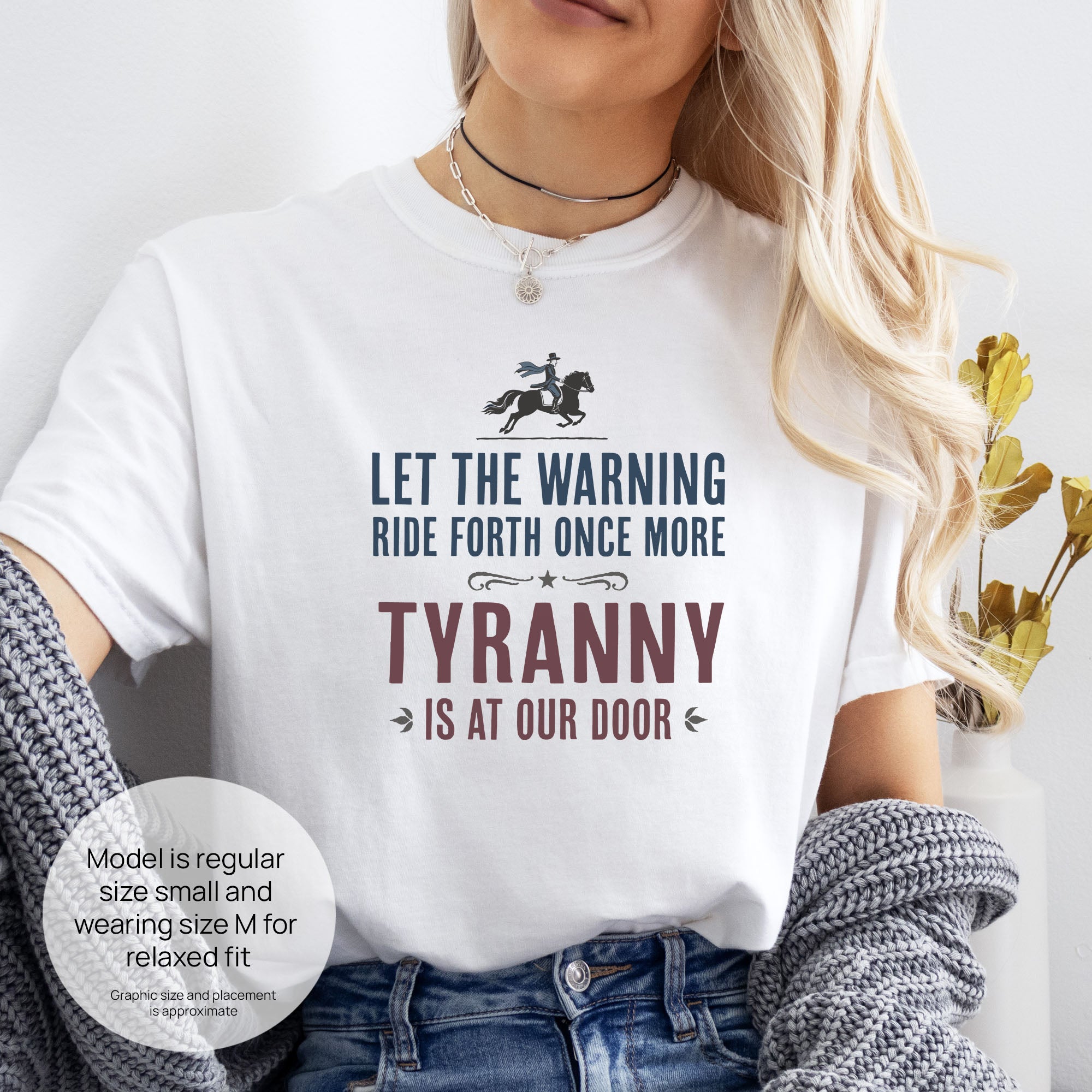 Female model wearing white t-shirt with a silhouette of Paul Revere on horseback and the text “Let the warning ride forth once more – Tyranny is at our door” in blue and red. Includes size and fit information for the model who is regular size small, and wearing a size M shirt for a relaxed fit.
