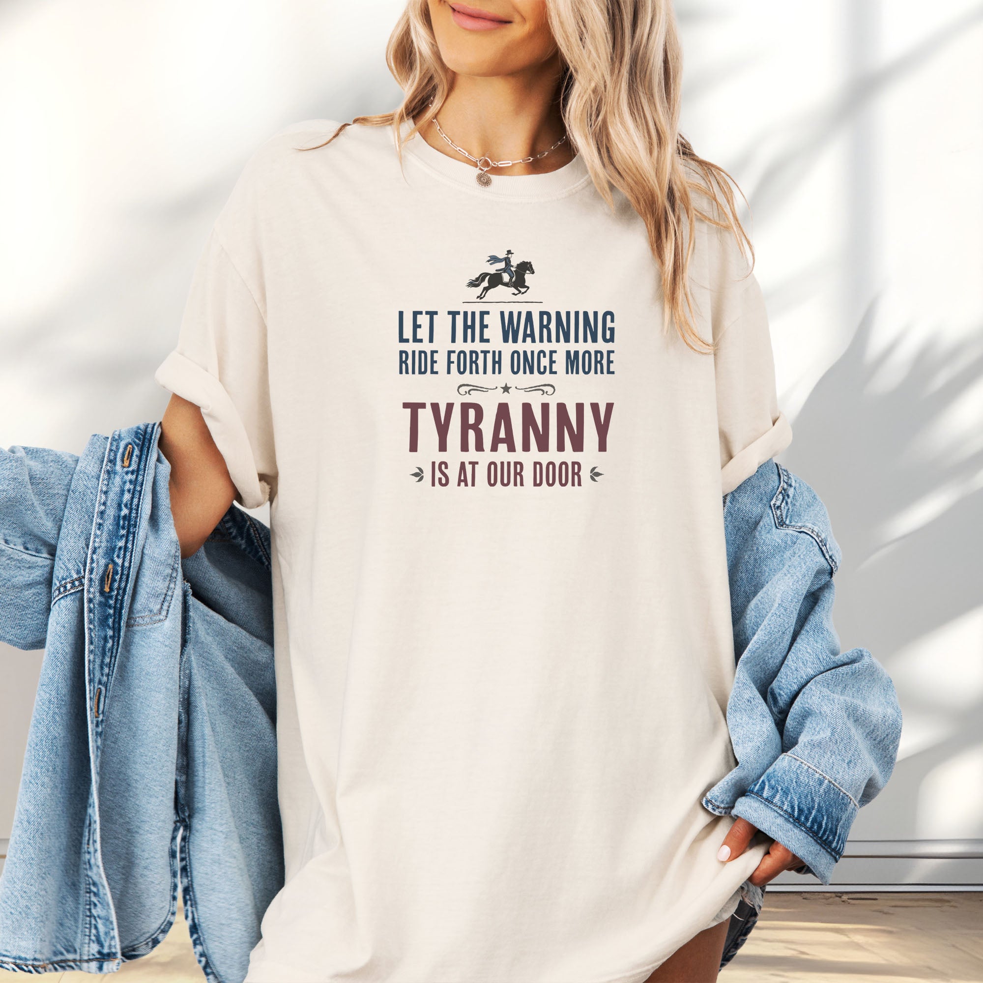 Female model wearing oversized ivory t-shirt with a silhouette of Paul Revere on horseback and the text “Let the warning ride forth once more – Tyranny is at our door” in blue and red.