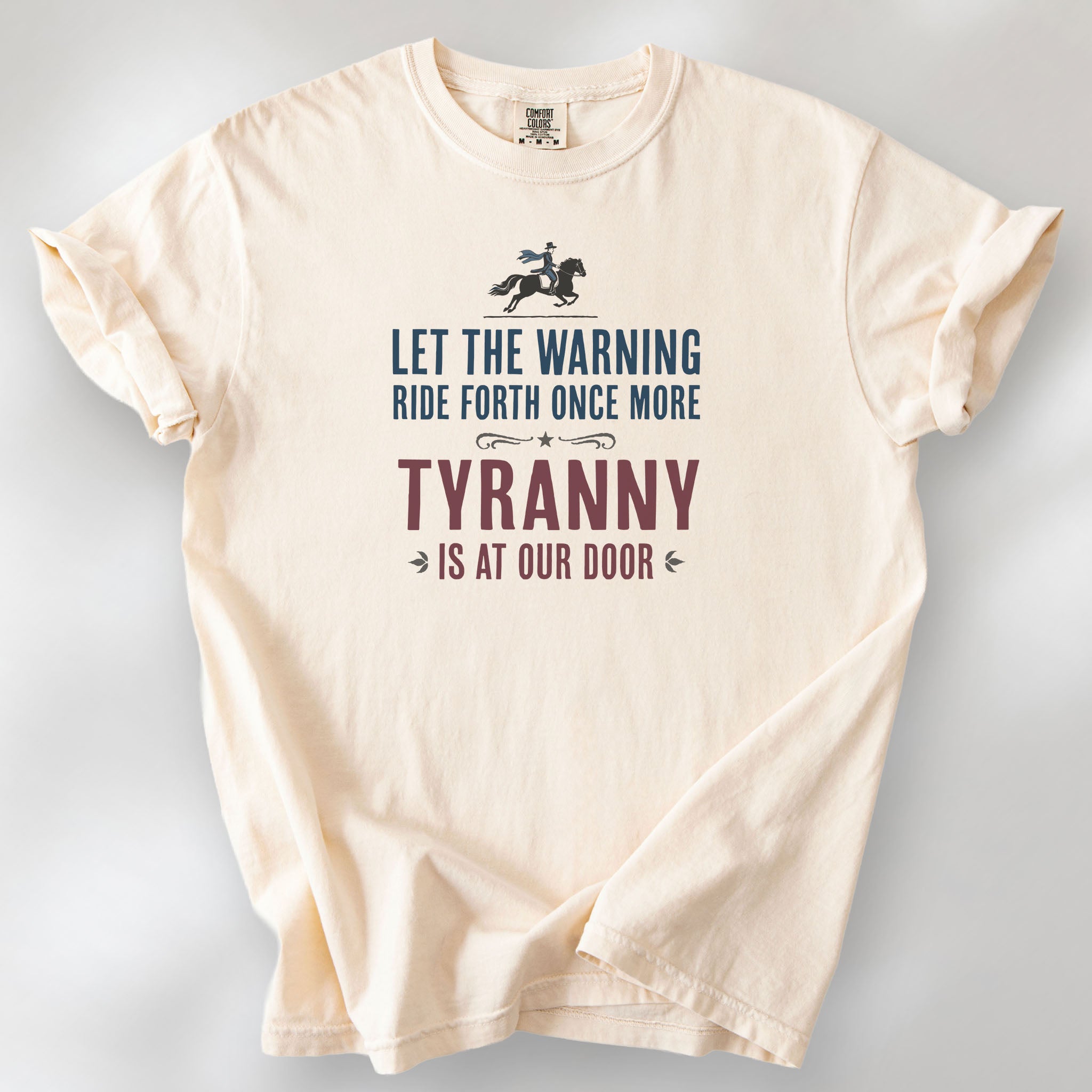 Ivory t-shirt with a silhouette of Paul Revere on horseback and the text “Let the warning ride forth once more – Tyranny is at our door” in blue and red.