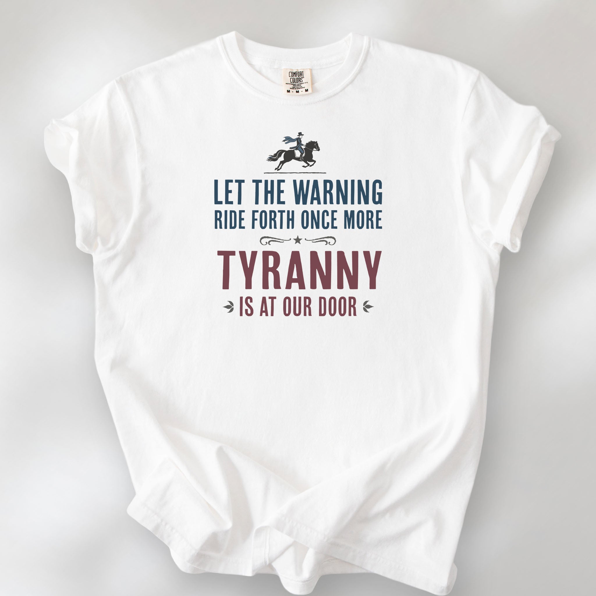 White t-shirt with a silhouette of Paul Revere on horseback and the text “Let the warning ride forth once more – Tyranny is at our door” in blue and red.
