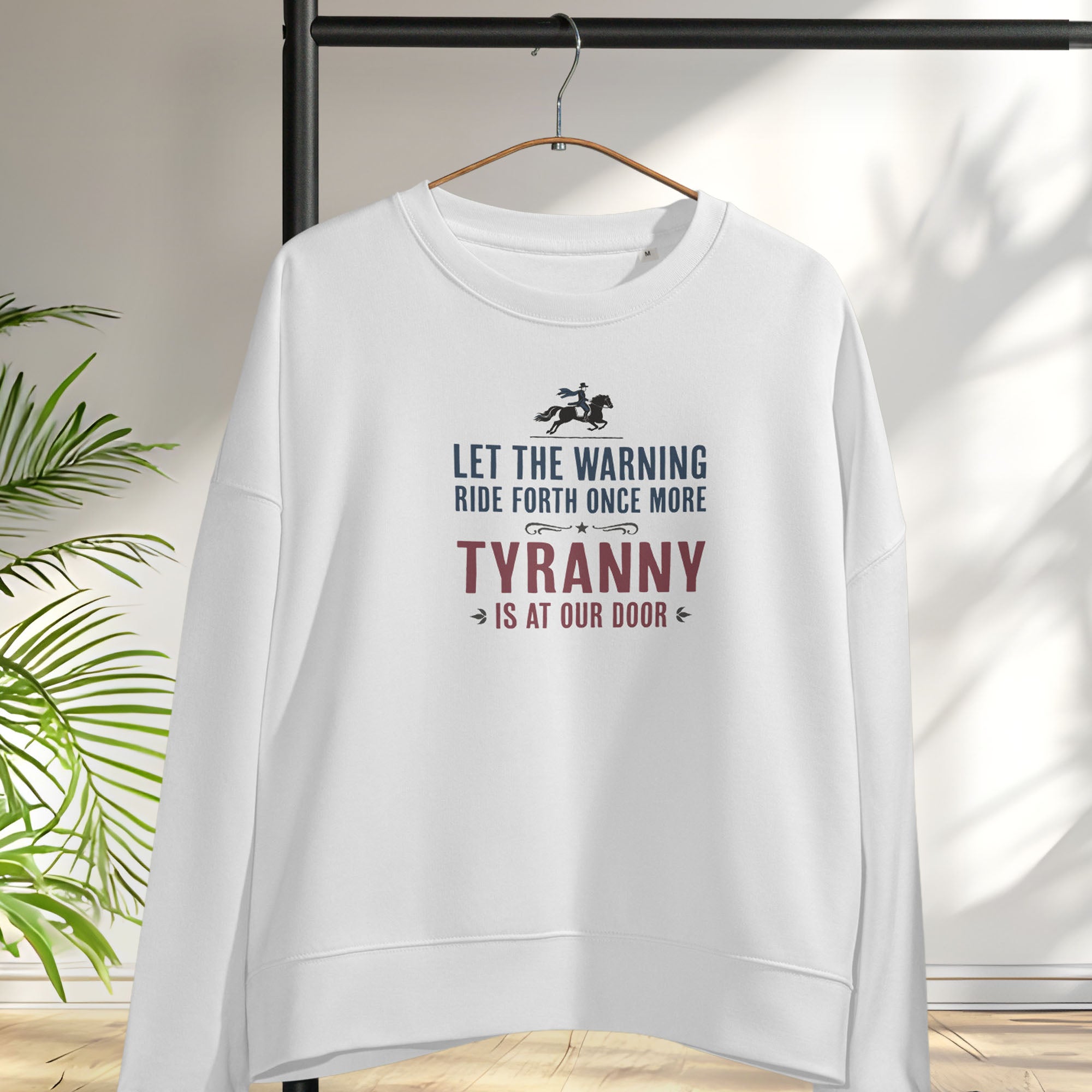 White sweatshirt with a silhouette of Paul Revere on horseback and the text “Let the warning ride forth once more – Tyranny is at our door” in blue and red, hanging on a hanger.