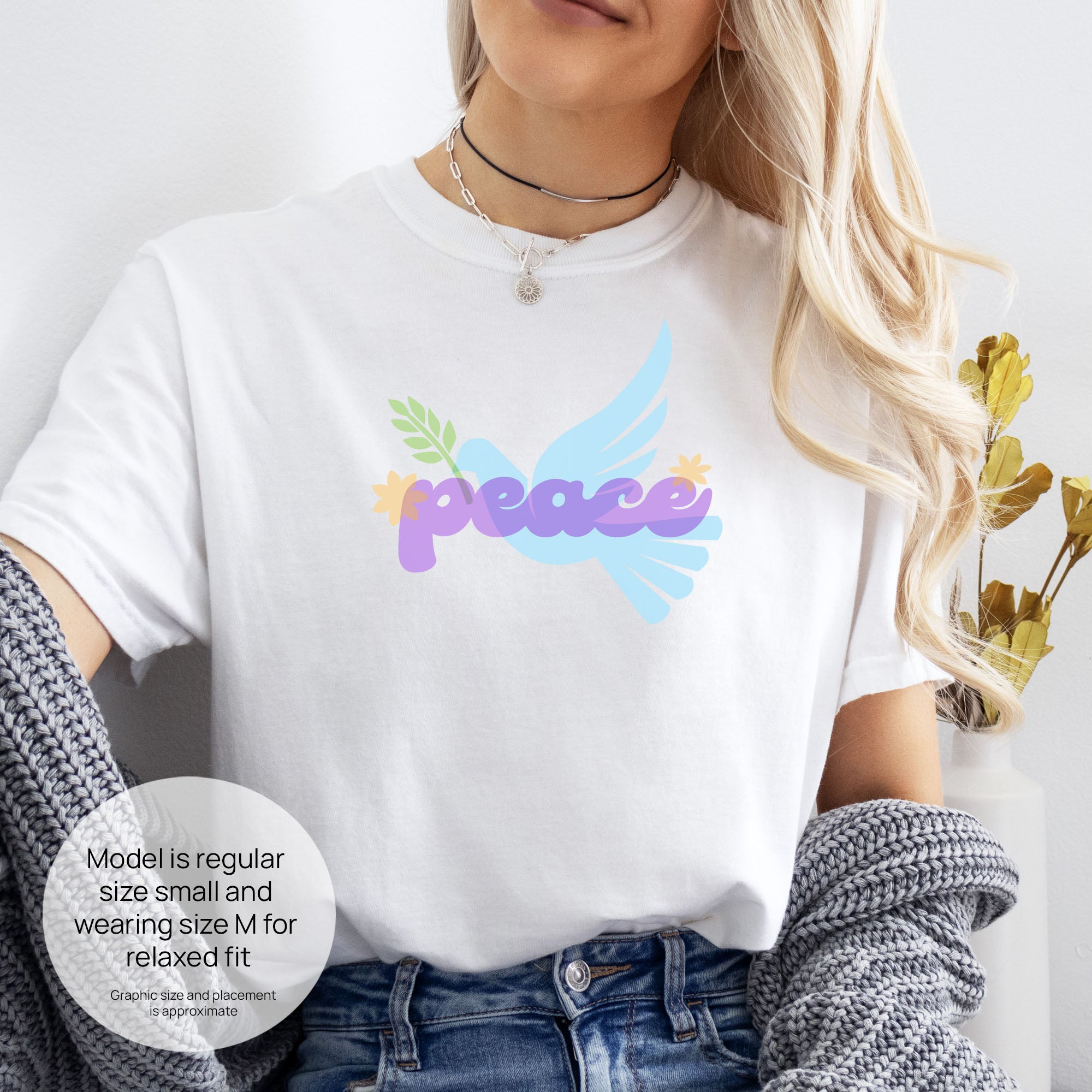 Female model wearing white t-shirt featuring a colorful retro peace dove carrying an olive branch with the word "peace" in groovy 1970s-style lettering.  Includes size and fit information for the model who is regular size small, and wearing a size M shirt for a relaxed fit.
