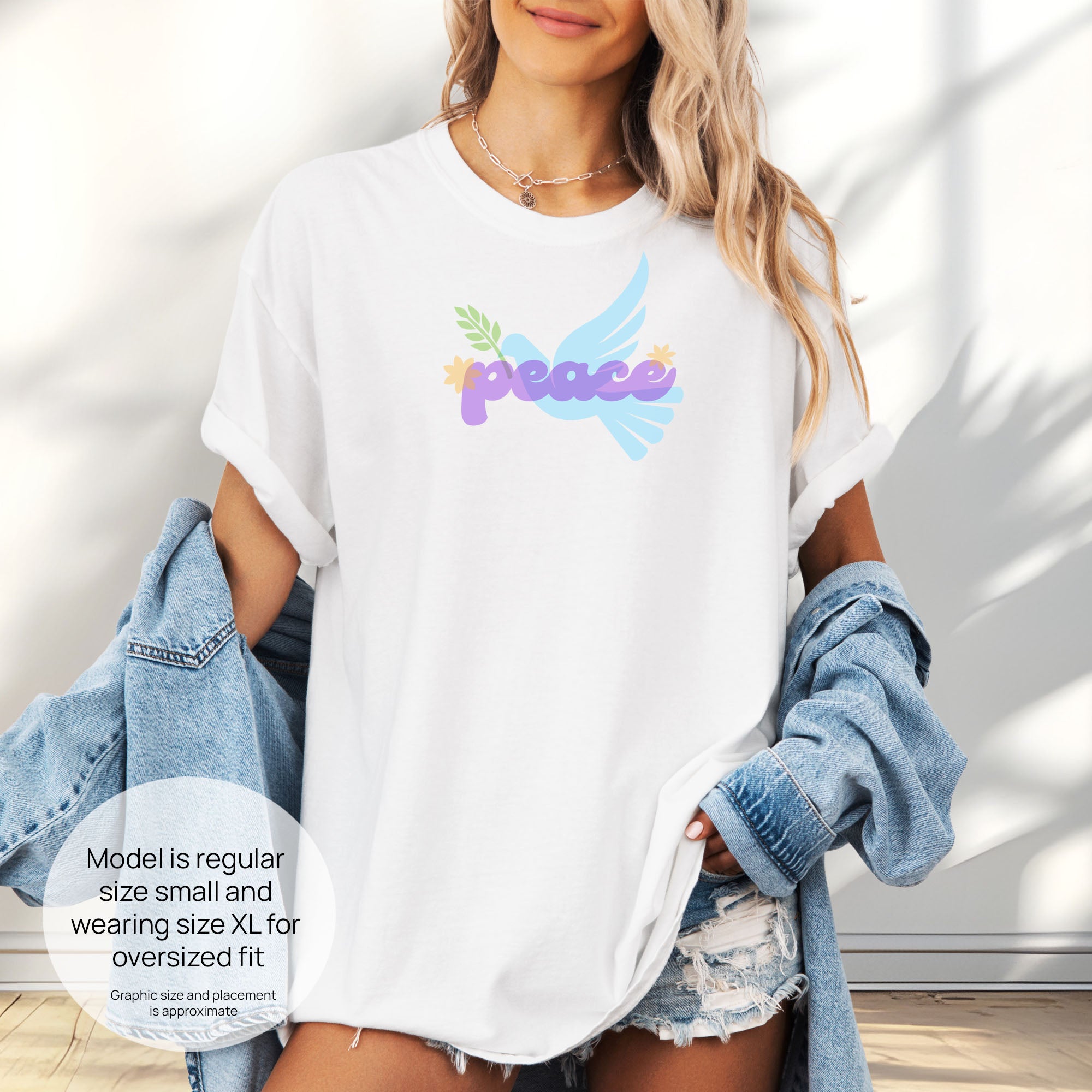 Female model wearing oversized white t-shirt featuring a colorful retro peace dove carrying an olive branch with the word "peace" in groovy 1970s-style lettering.  Includes size and fit information for the model who is regular size small, and wearing a size XL shirt for an oversized fit.
