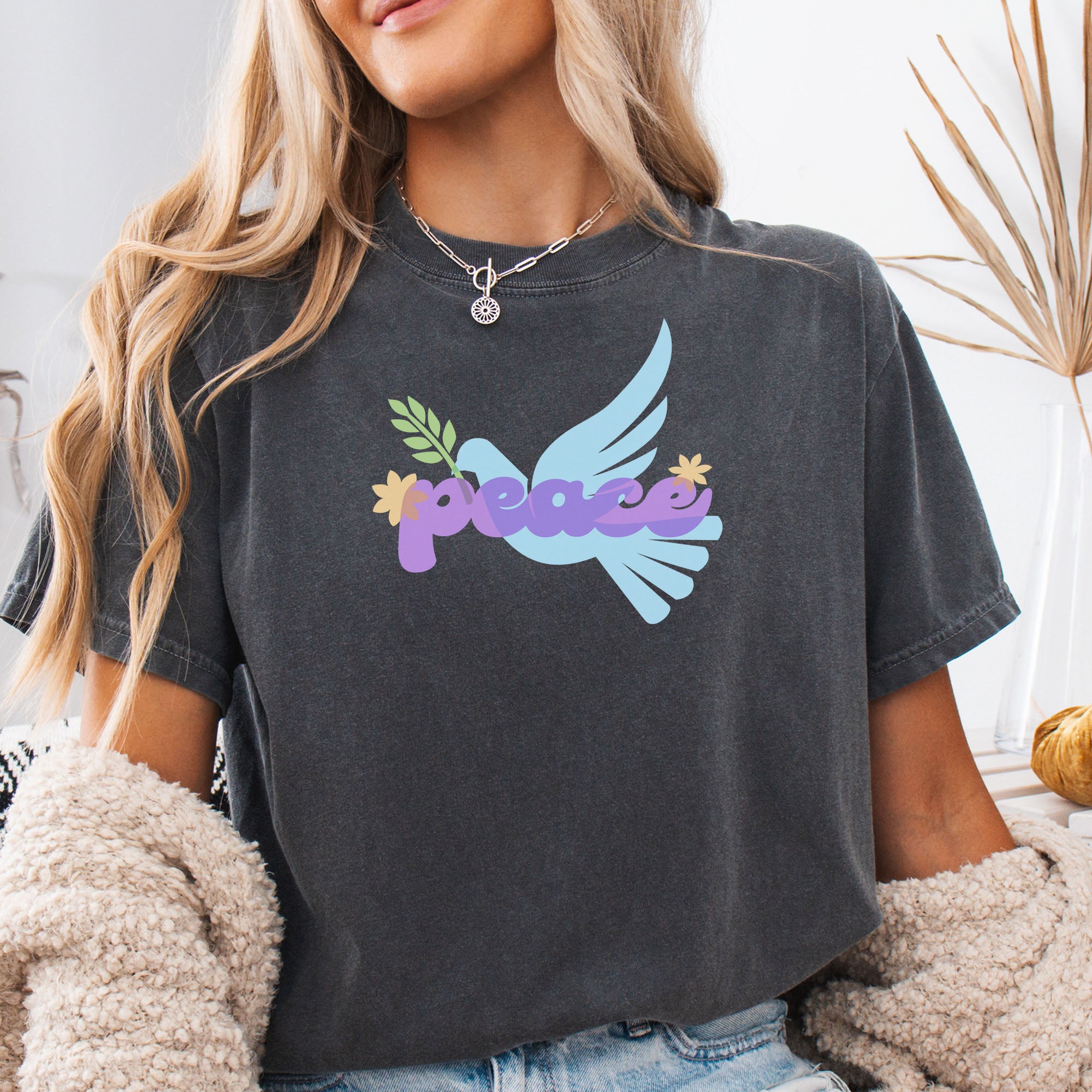 Female model wearing pepper t-shirt featuring a colorful retro peace dove carrying an olive branch with the word "peace" in groovy 1970s-style lettering. 