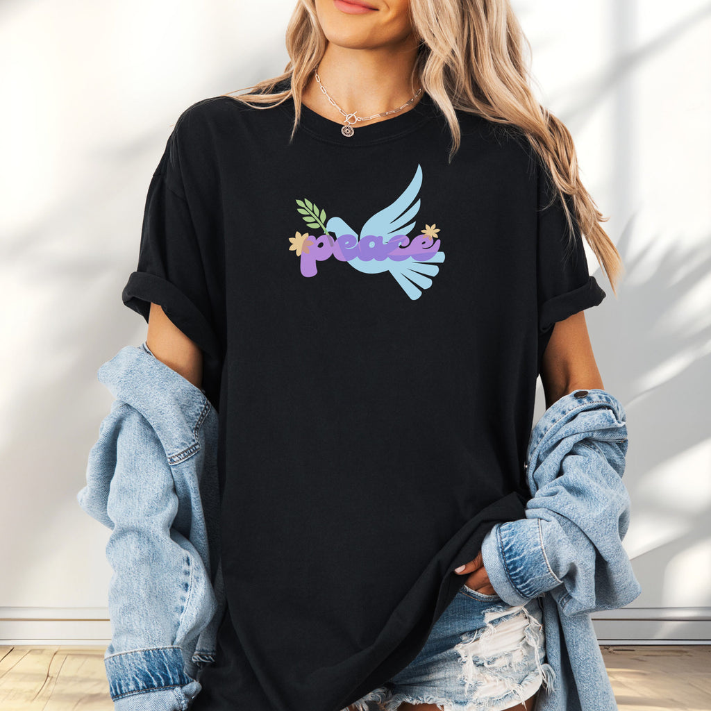 Female model wearing oversized black t-shirt featuring a colorful retro peace dove carrying an olive branch with the word "peace" in groovy 1970s-style lettering. 