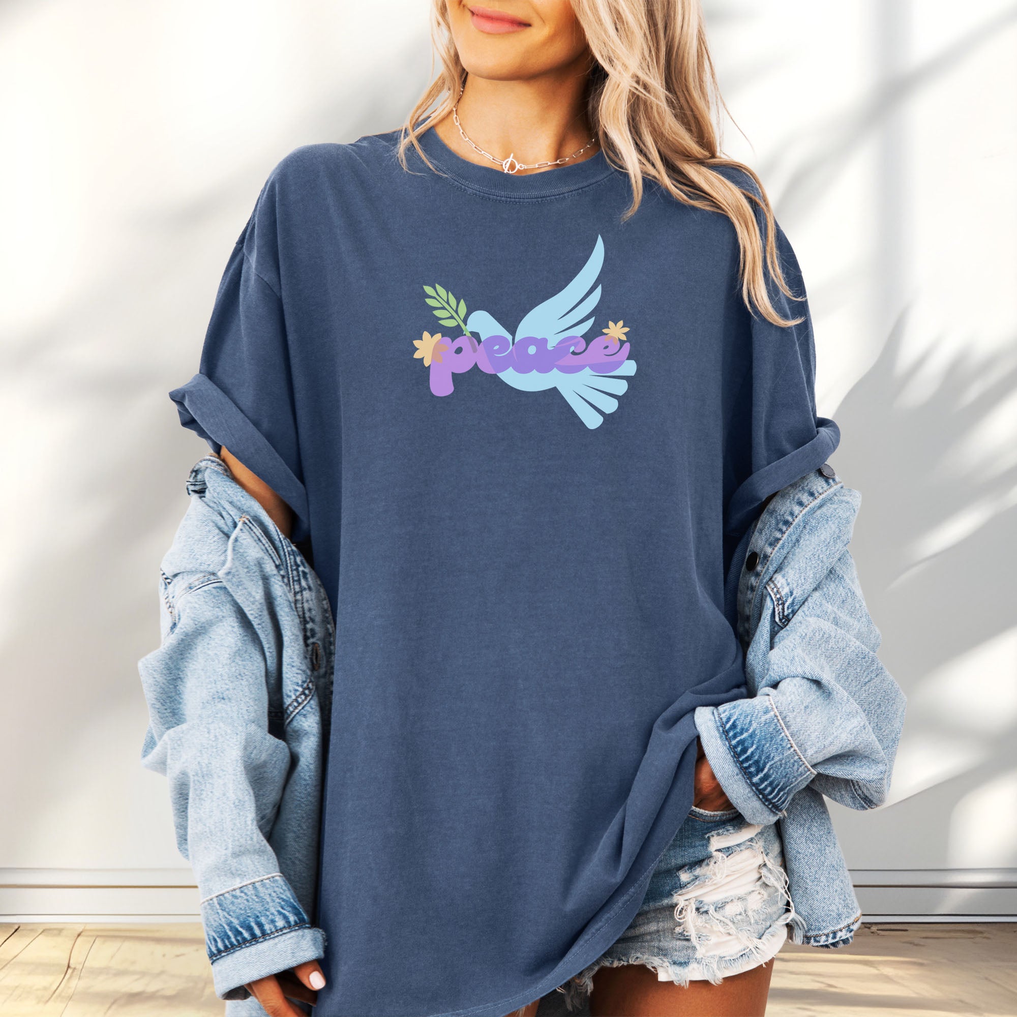 Female model wearing oversized denim t-shirt featuring a colorful retro peace dove carrying an olive branch with the word "peace" in groovy 1970s-style lettering. 