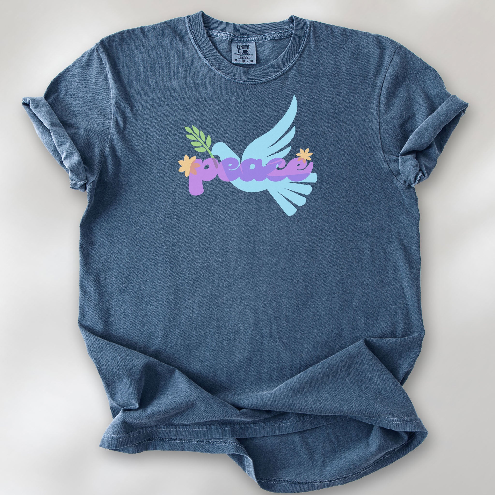 Denim t-shirt featuring a colorful retro peace dove carrying an olive branch with the word "peace" in groovy 1970s-style lettering. 
