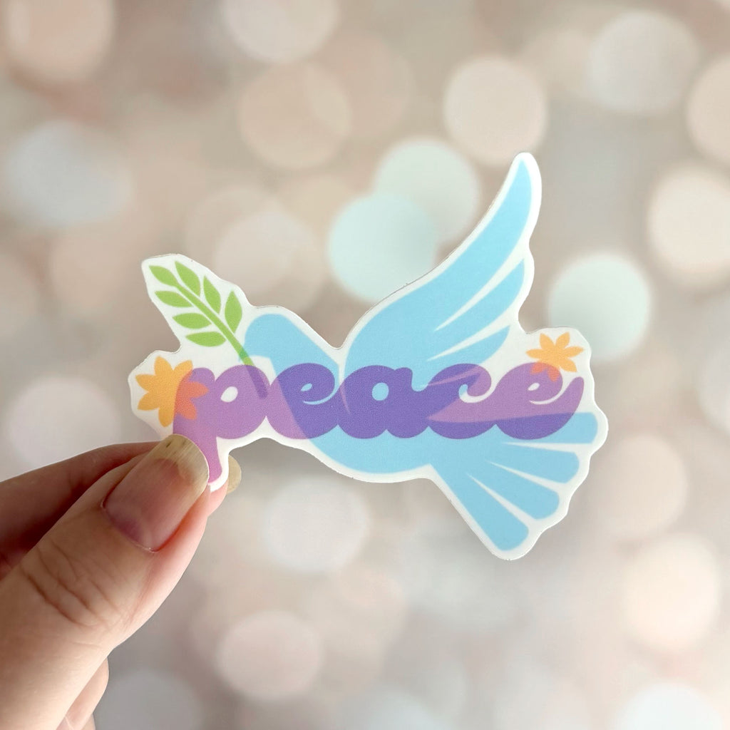 Blue peace dove vinyl sticker with olive branch and purple "peace" lettering, shown held in hand against a soft neutral bokeh background.