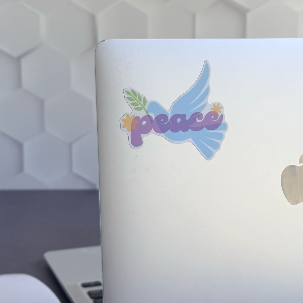 Blue peace dove vinyl sticker with olive branch and purple "peace" lettering, shown stuck on a silver apple laptop computer.