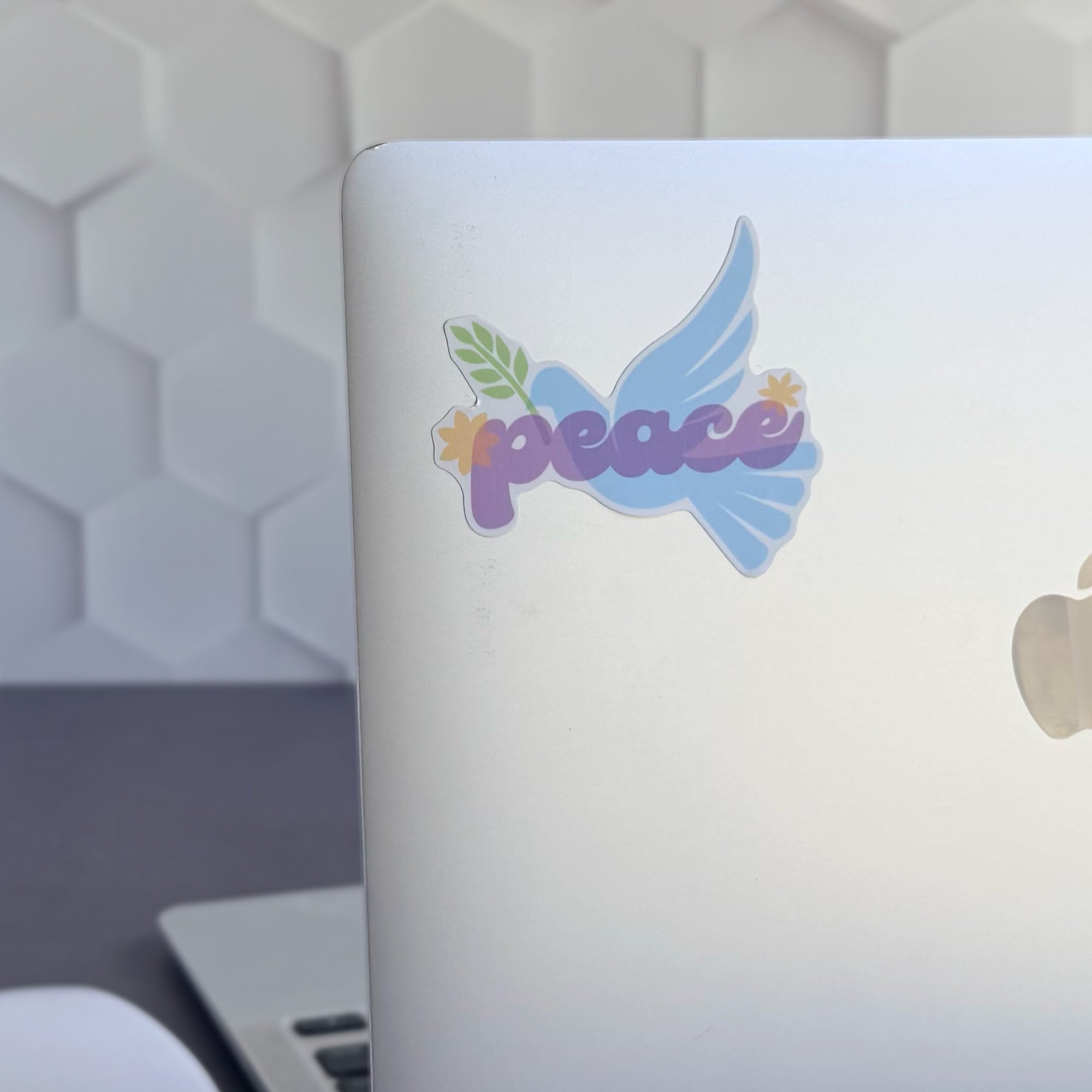 Blue peace dove vinyl sticker with olive branch and purple "peace" lettering, shown stuck on a silver apple laptop computer.