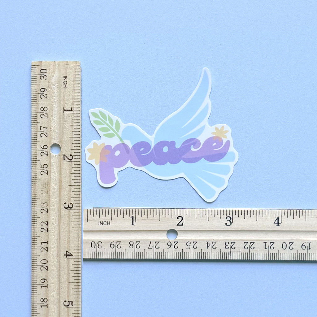 Blue peace dove vinyl sticker with olive branch and purple "peace" lettering, shown with two rulers to demonstrate approximate size of 3 inches by 3 inches.