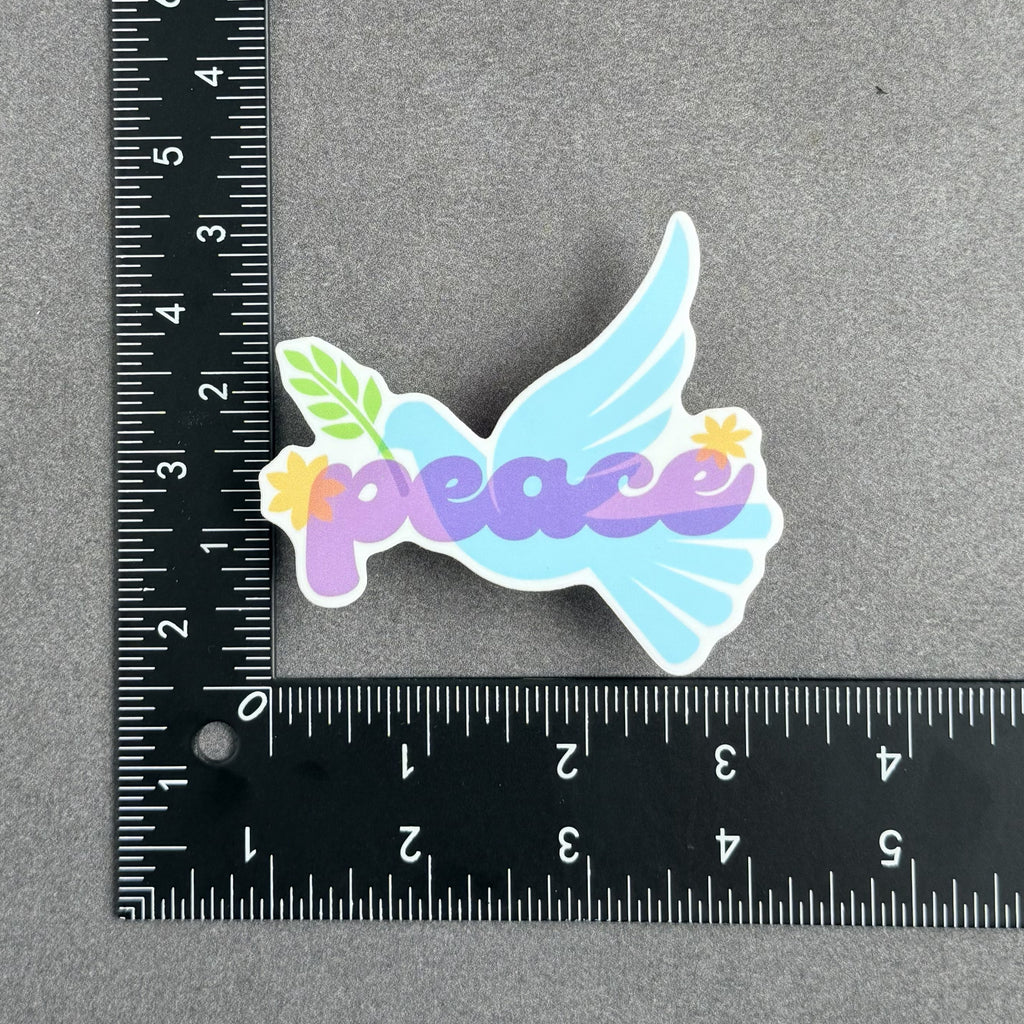 Blue peace dove vinyl sticker with olive branch and purple "peace" lettering, shown with ruler to demonstrate approximate size of 3 inches by 3 inches.