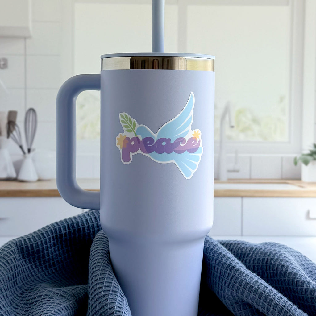 Blue peace dove vinyl sticker with olive branch and purple "peace" lettering, stuck on a tumbler in a kitchen.