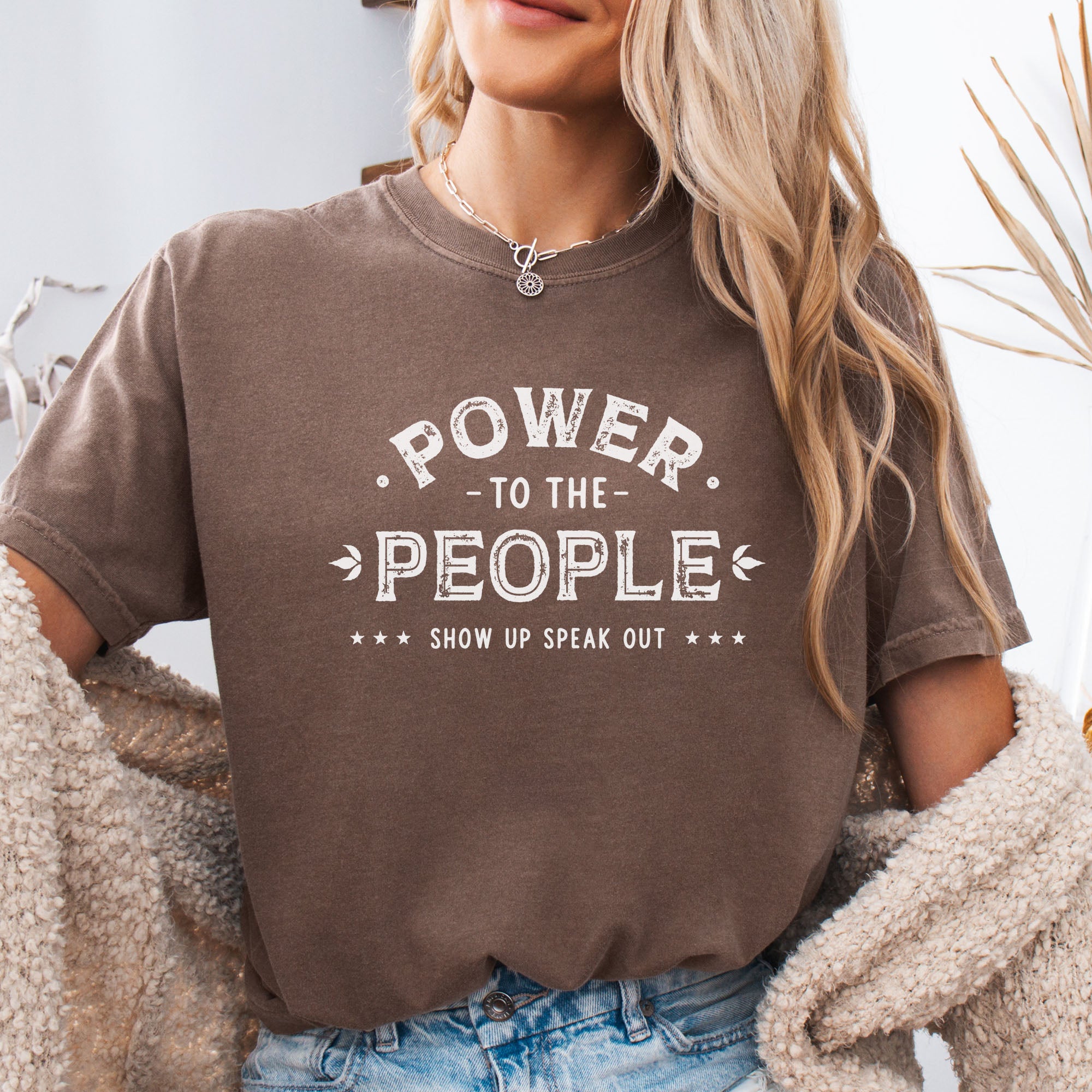 Female model wearing espresso t-shirt with vintage-style distressed text reading “Power to the People — Show Up Speak Out”.