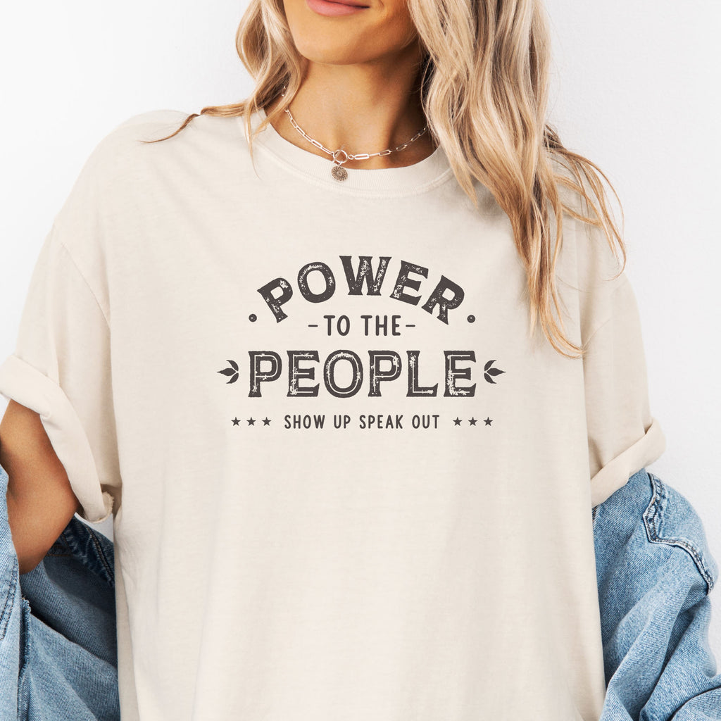 Female model wearing oversized ivory t-shirt with vintage-style distressed text reading “Power to the People — Show Up Speak Out”.
