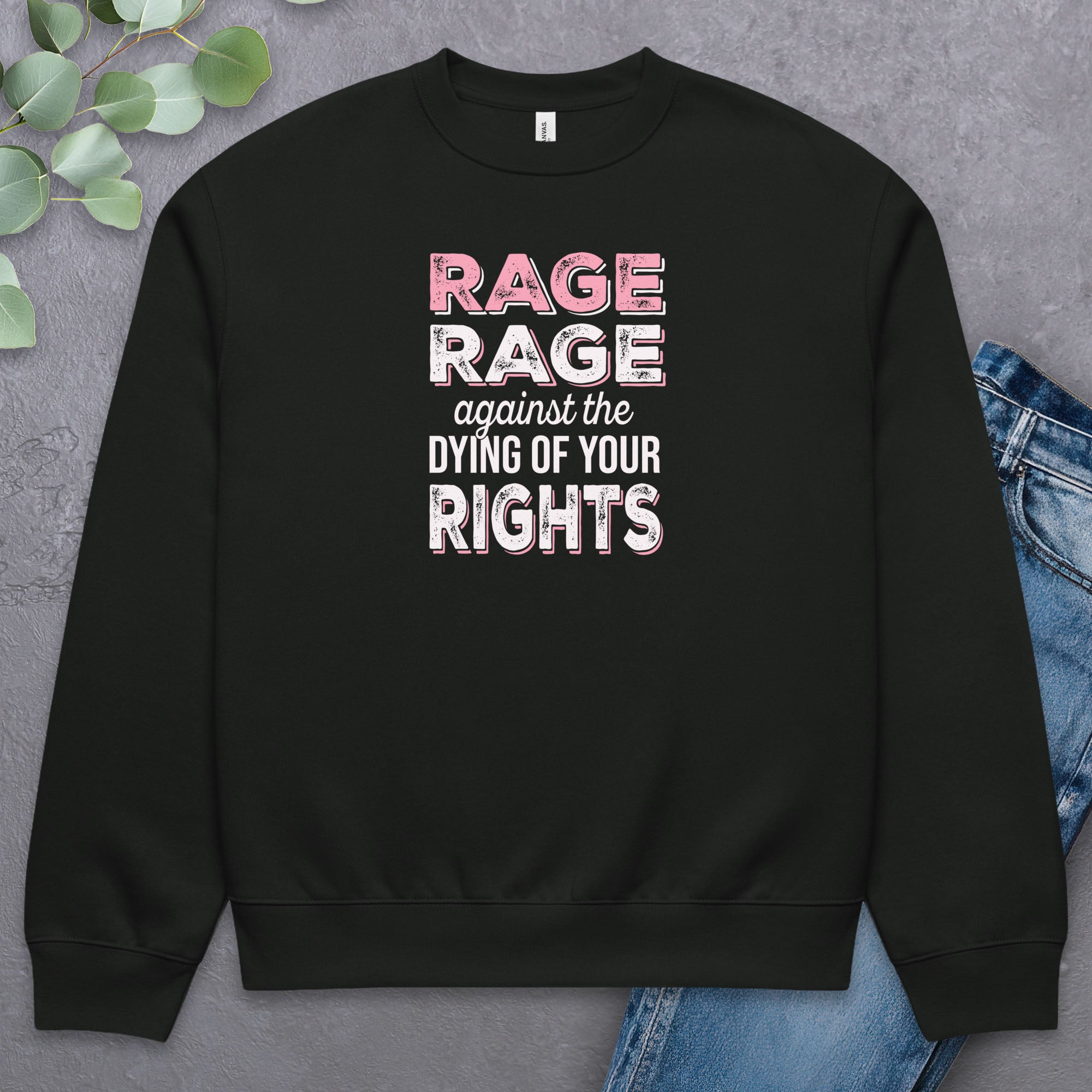Black protest sweatshirt with bold pink and white text reading “rage rage against the dying of your rights” – feminist anti-trump activist shirt. Laying flat with a pair of jeans.