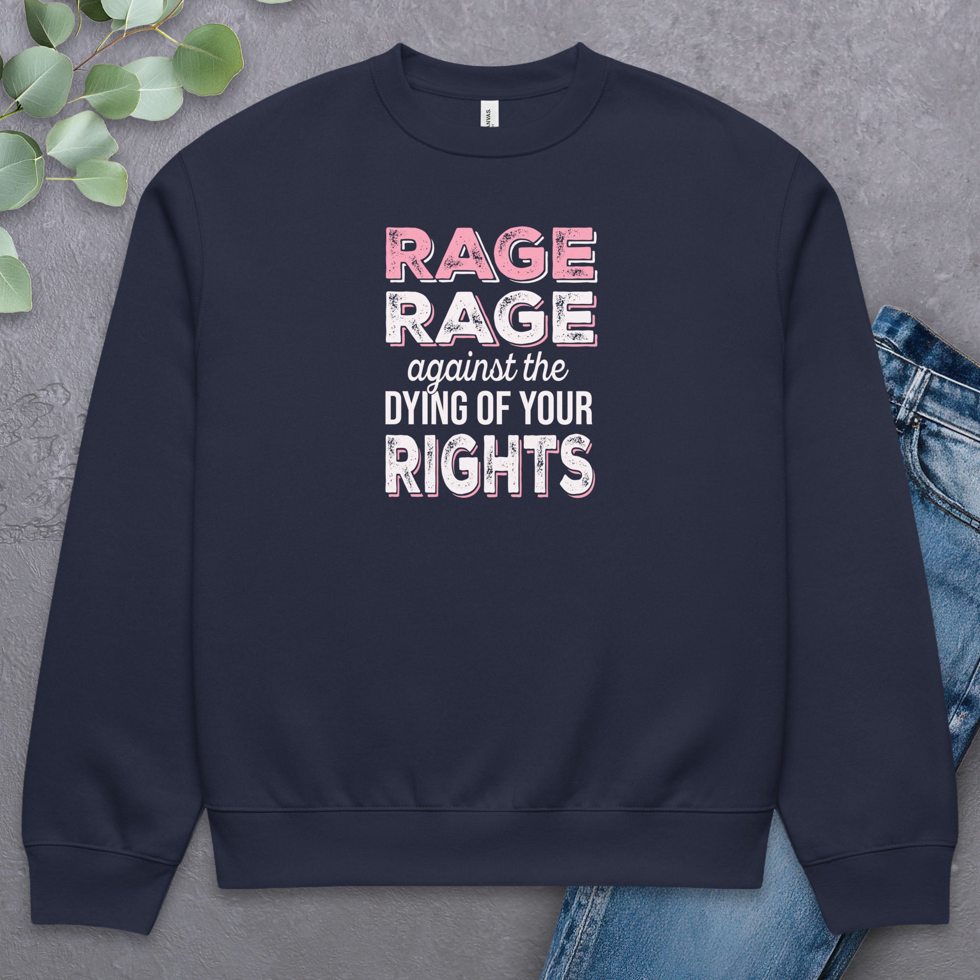 Navy protest sweatshirt with bold pink and white text reading “rage rage against the dying of your rights” – feminist anti-trump activist shirt. Laying flat with a pair of jeans.
