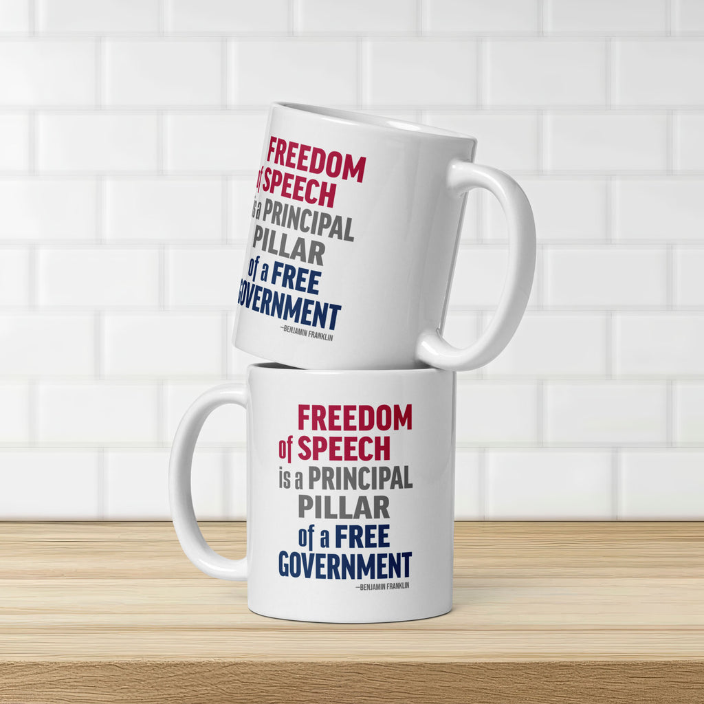 Two white ceramic mugs with bold patriotic red, blue, and grey text that reads "Freedom of speech is a principal pillar of a free government. – Ben Franklin." Mugs are stacked on a counter in front of white subway tile.