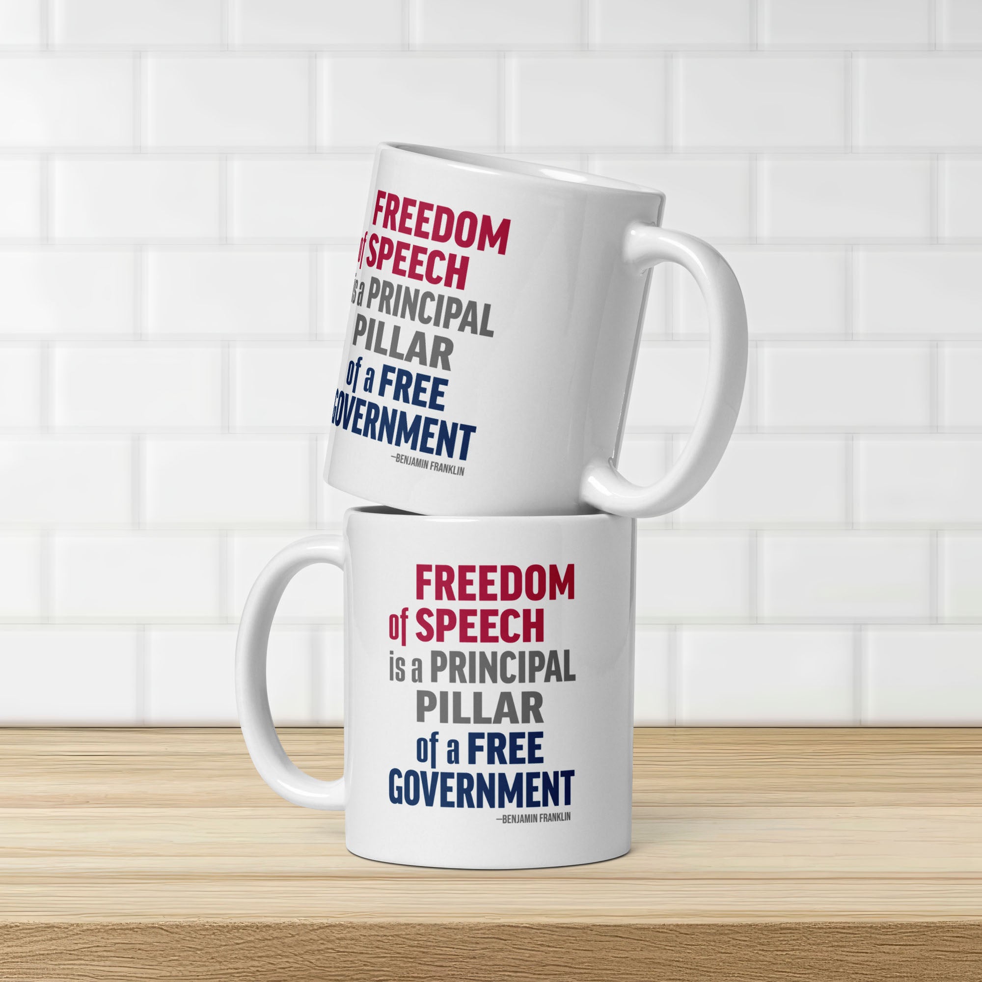 Two white ceramic mugs with bold patriotic red, blue, and grey text that reads "Freedom of speech is a principal pillar of a free government. – Ben Franklin." Mugs are stacked on a counter in front of white subway tile.